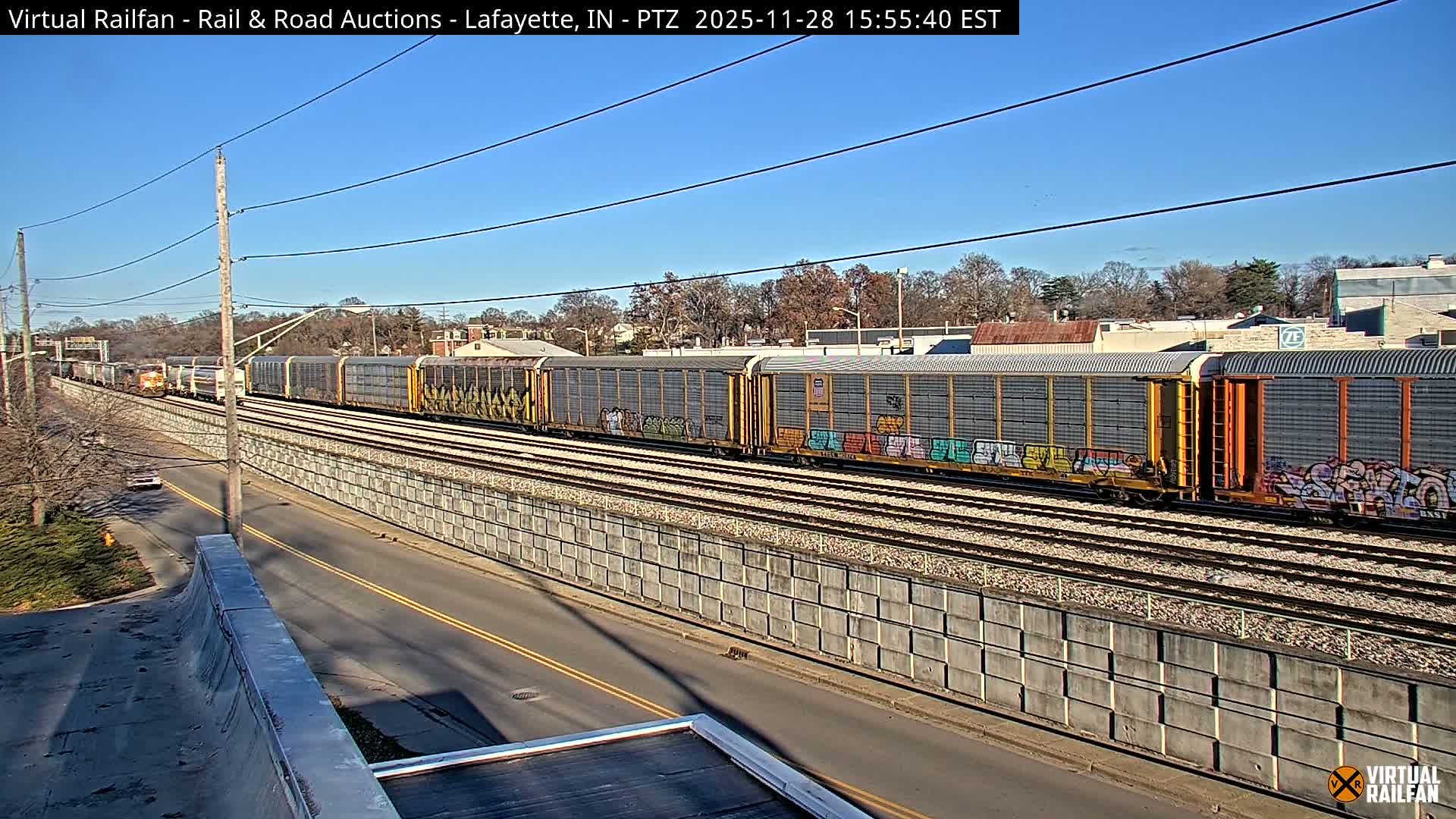 A long freight train with numerous cars, some displaying colorful graffiti, travels on multiple tracks alongside a road and concrete retaining wall, beneath a clear blue sky in an urban-suburban landscape with bare trees and distant buildings.
