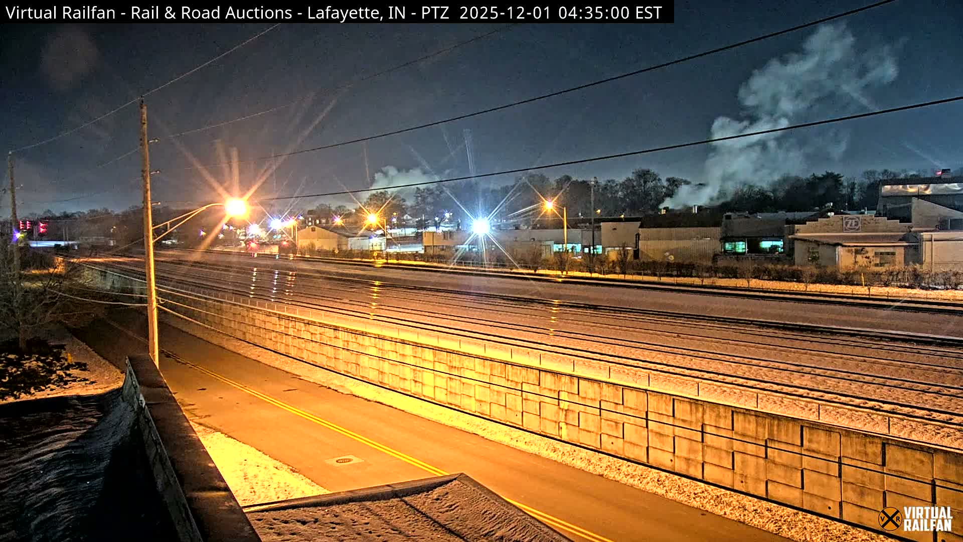 Multiple parallel railroad tracks and an illuminated road run through an urban landscape covered in light snow at night, with bright streetlights and plumes of steam rising from distant buildings.
