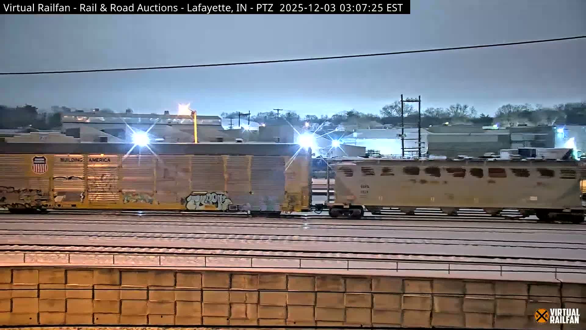Two freight train cars, one yellow and graffitied and another gray, are illuminated by bright artificial lights as they sit on multiple snow-covered tracks outdoors during an overcast winter night, with industrial buildings in the background.