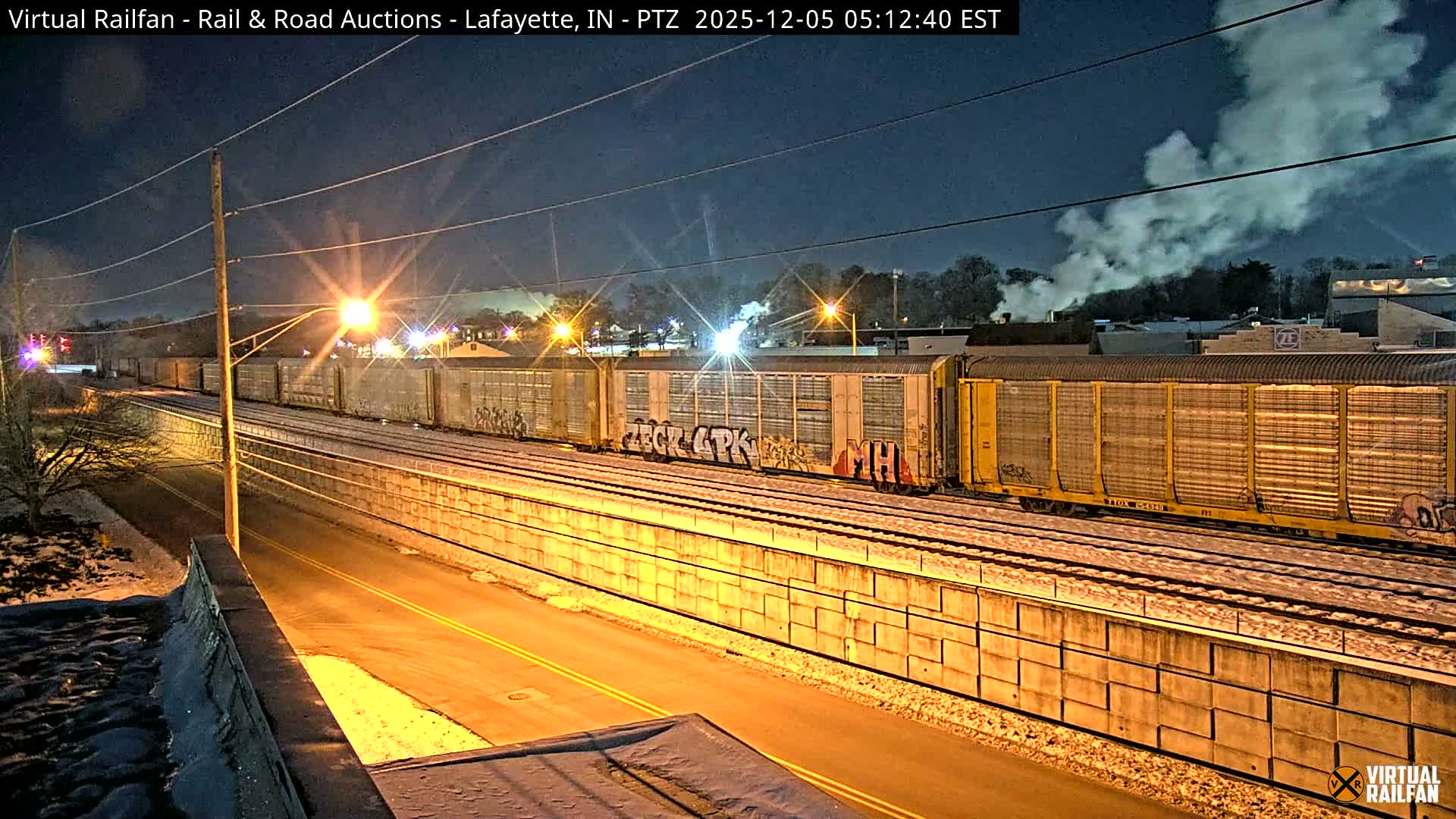A long freight train with graffiti on some cars sits on snowy tracks beside a brightly lit road and utility pole under a dark, clear early morning sky, with steam plumes rising from distant buildings, indicating cold weather.