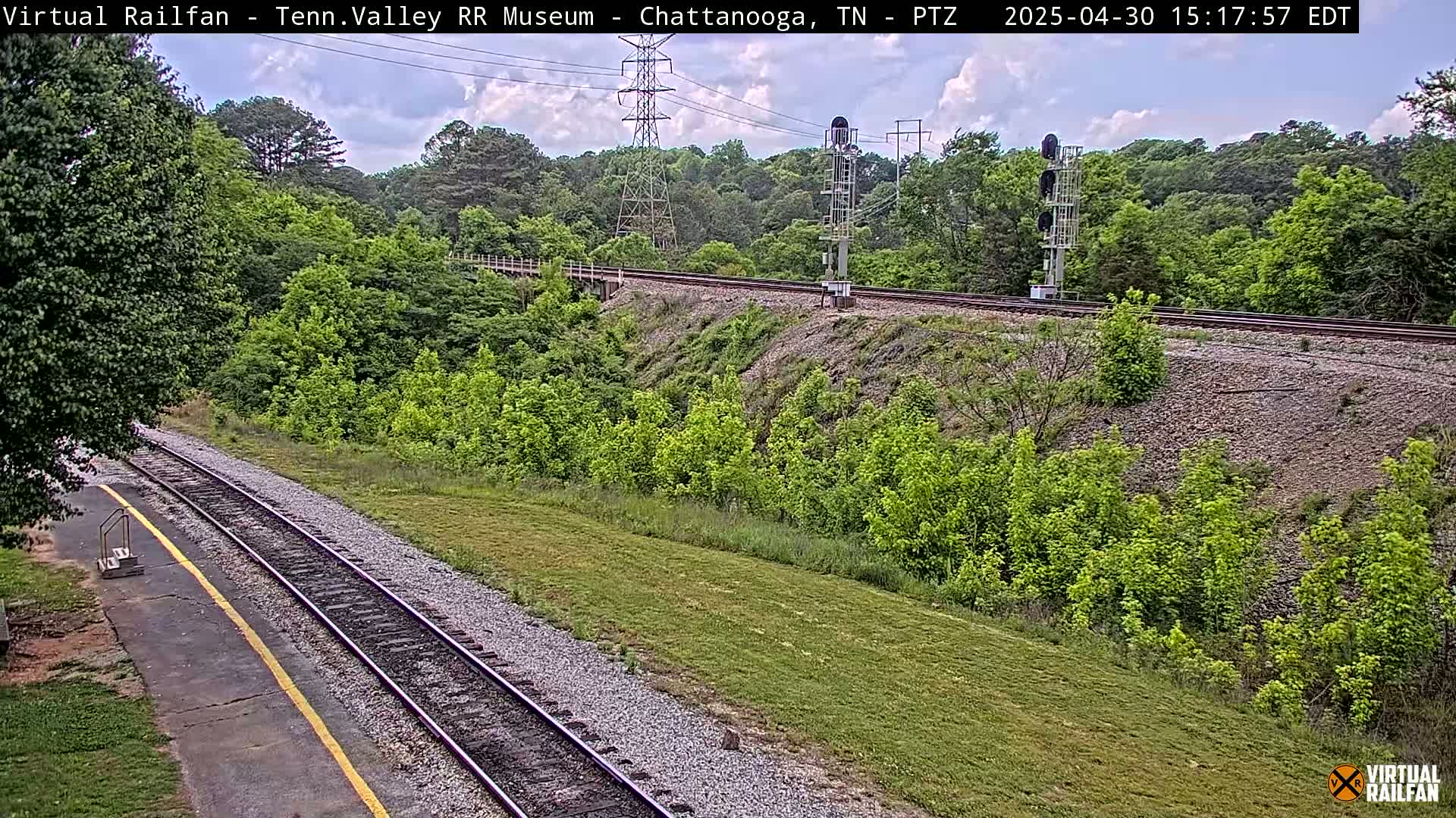 A mostly sunny day shows a train track curving through a lush green landscape, with a small train platform visible in the foreground.