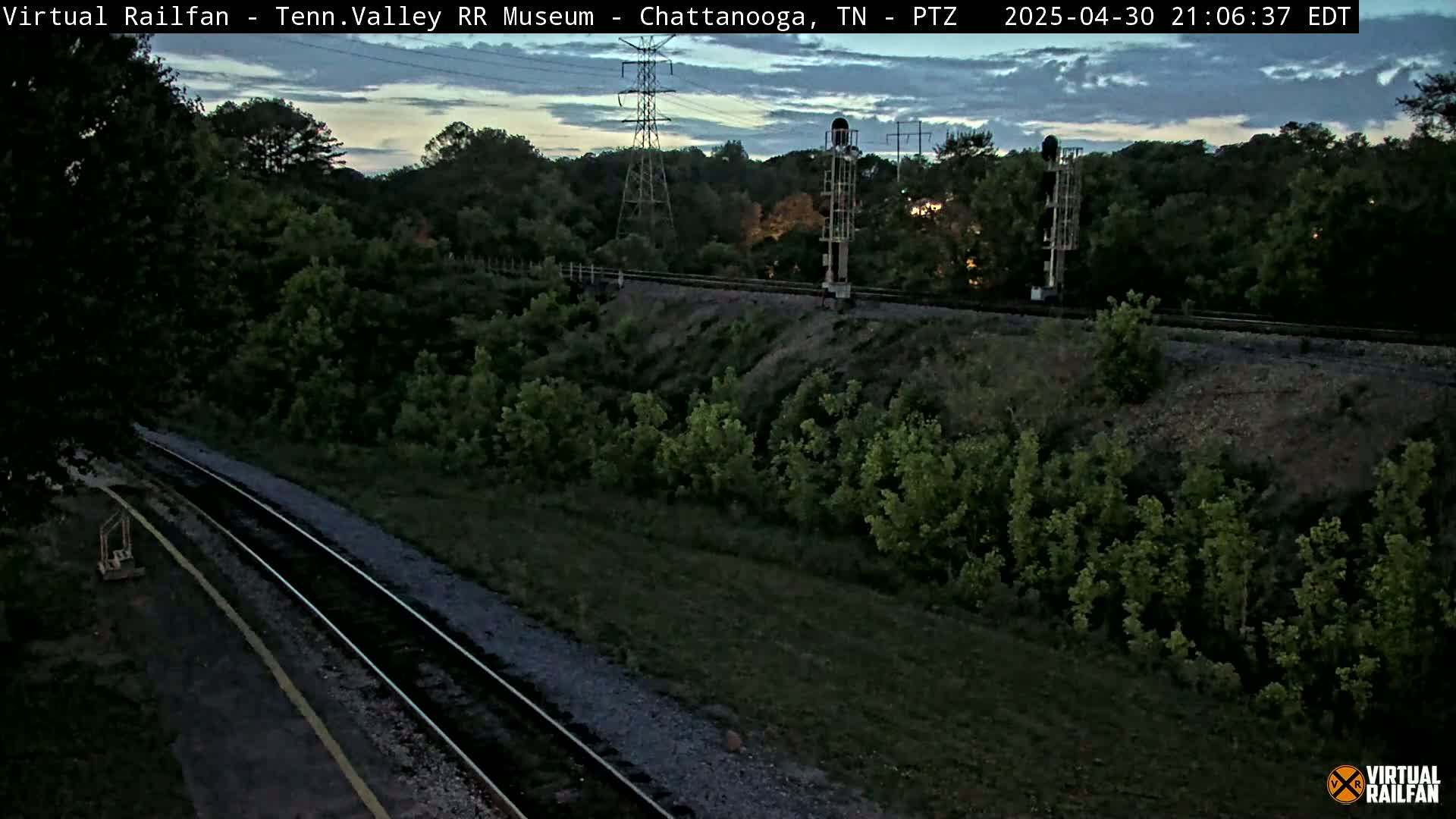Under a twilight sky, a train track winds through a lush green landscape, passing near tall signal towers.