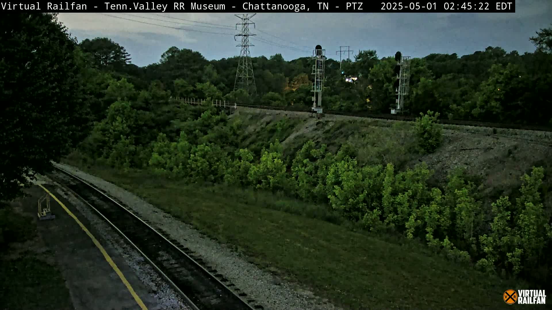A twilight view of railroad tracks winding through a lush green landscape, with power lines and signal equipment visible in the distance.
