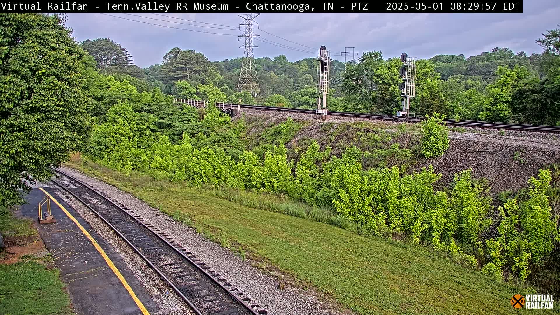 A mostly cloudy day shows a railway track winding through lush green vegetation, alongside a power line and signal lights.