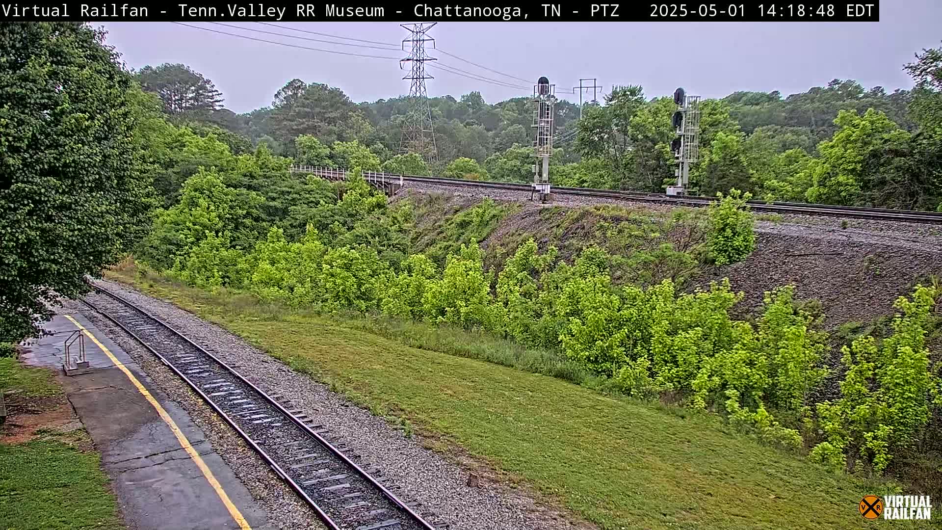 A high-angle view of a railway track cutting through a lush green landscape under an overcast sky.