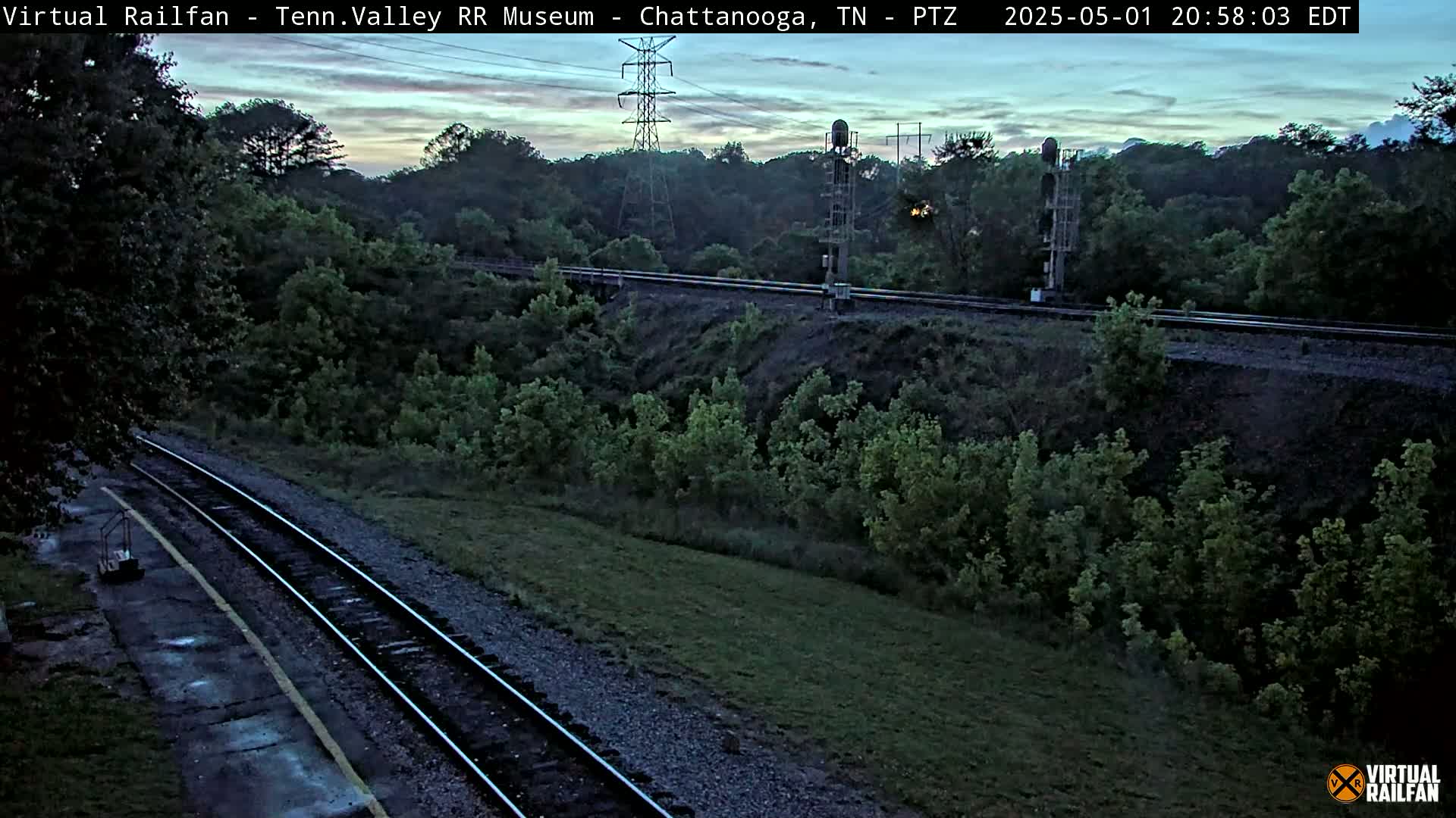 A twilight view of a train track running through a lush green landscape, with power lines and signal equipment visible in the background.