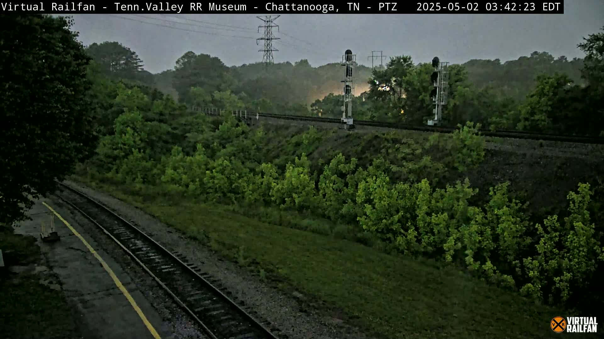 A wet, overcast scene shows a railroad track curving through lush green vegetation under a gray sky.