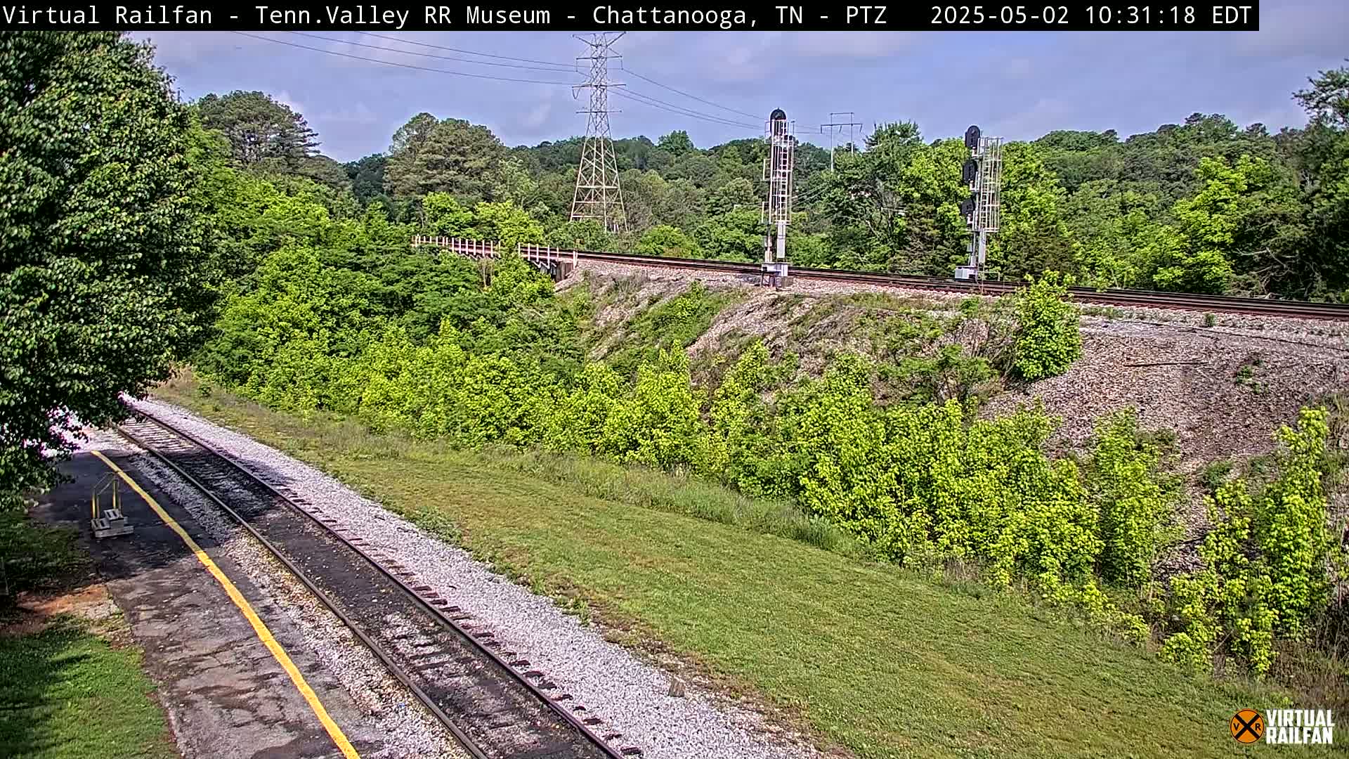 A mostly sunny view of railroad tracks winding through a lush green landscape, with a tall transmission tower visible in the background.
