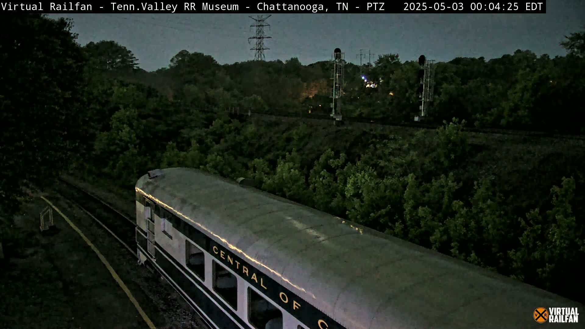 A dark gray passenger train car moves through a nighttime landscape of trees and railway infrastructure under a twilight sky.