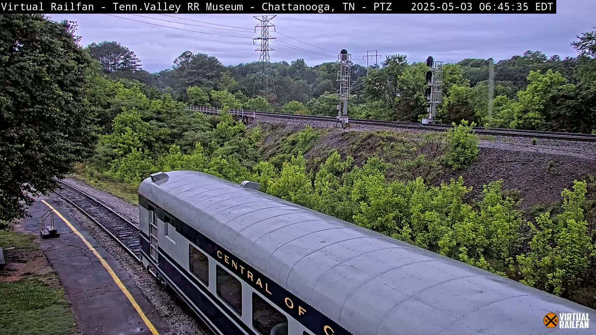 A silver passenger train car is visible on a railway track that runs through a lush green landscape under an overcast sky.