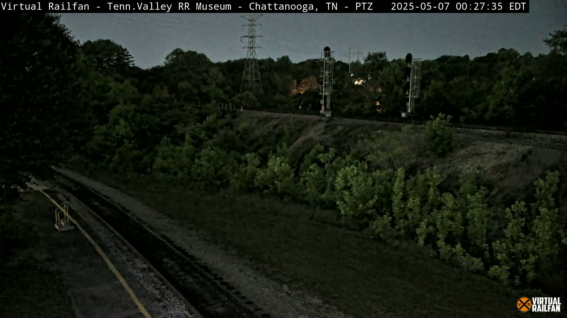 A nighttime view of train tracks surrounded by dark green vegetation under a twilight sky.