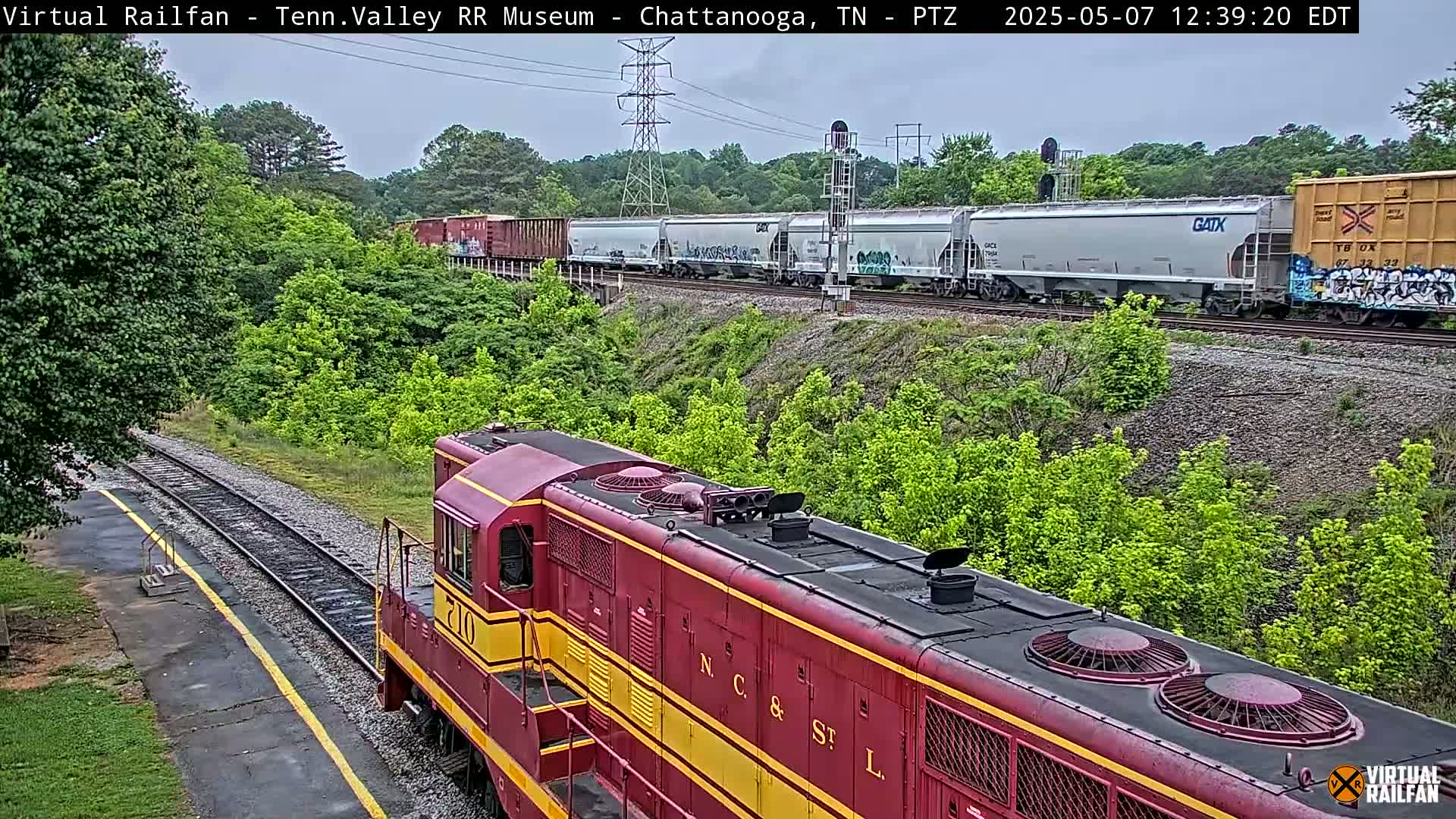 A red and yellow diesel locomotive is in the foreground, facing the viewer, on a railway track next to a lush green embankment, with a long freight train visible in the background under an overcast sky.