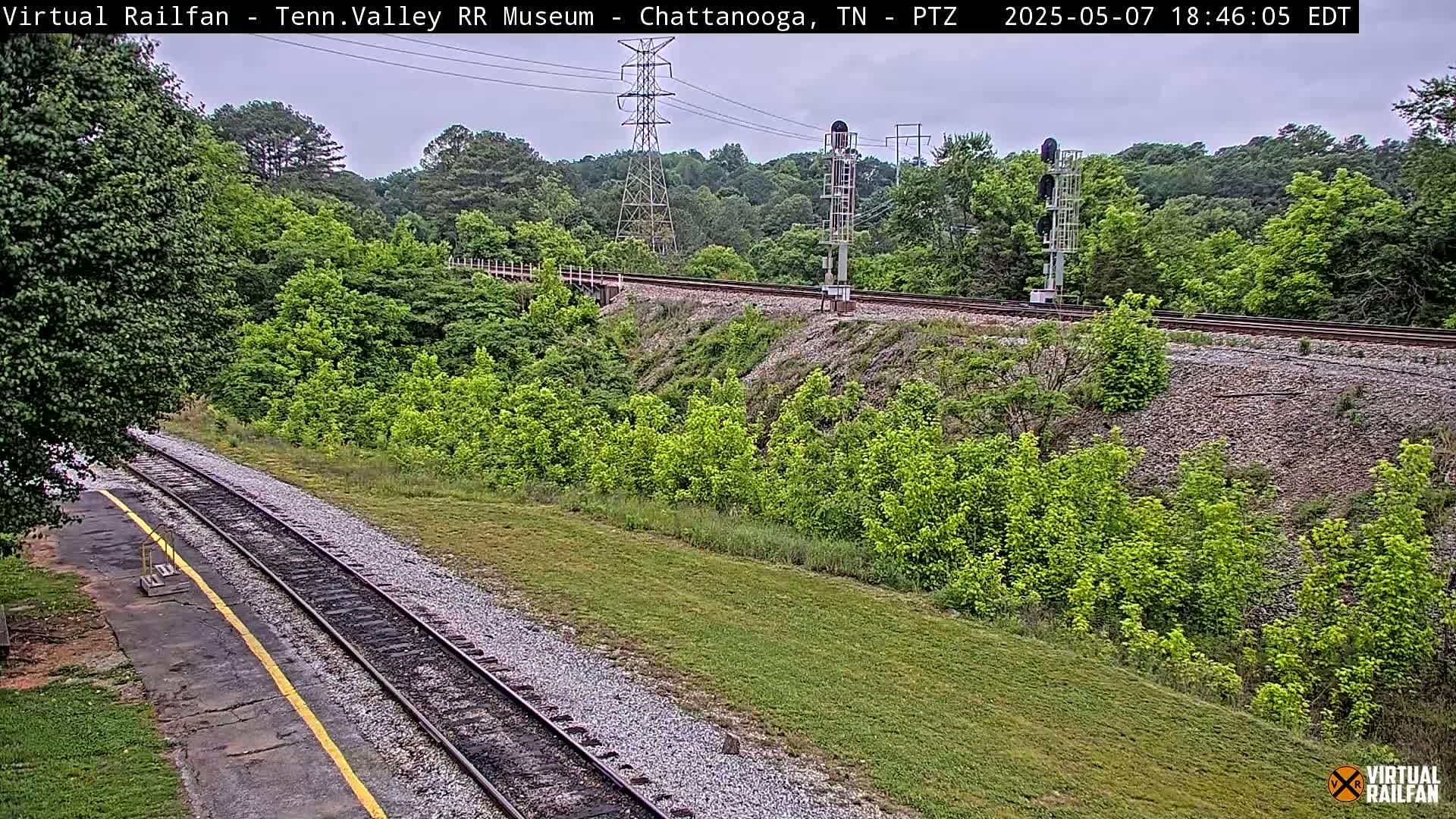 A mostly cloudy day shows a railroad track winding through a lush green landscape, with a tall power line and signal lights visible in the distance.