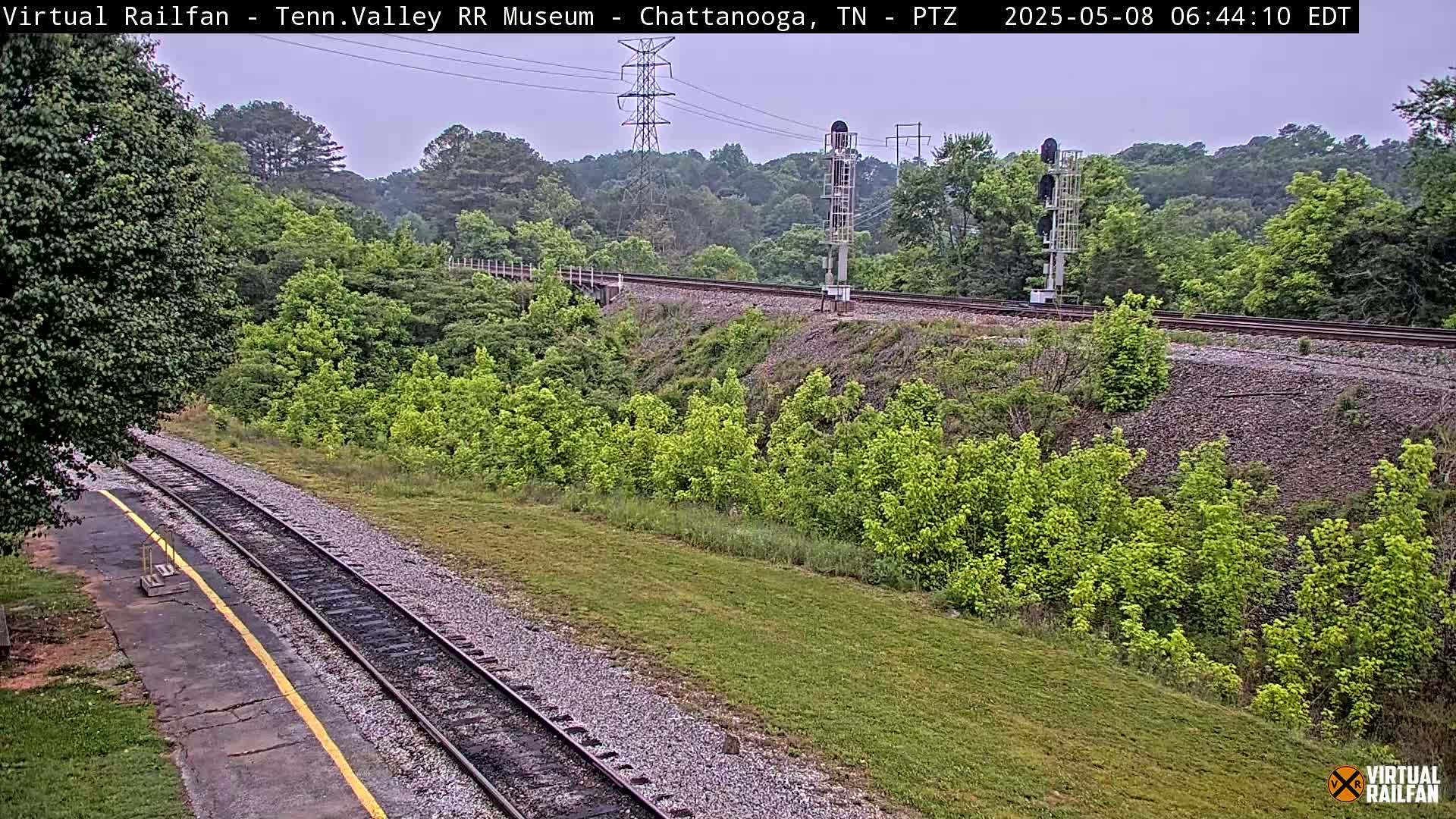 A section of railroad tracks runs through a lush green landscape under an overcast sky.