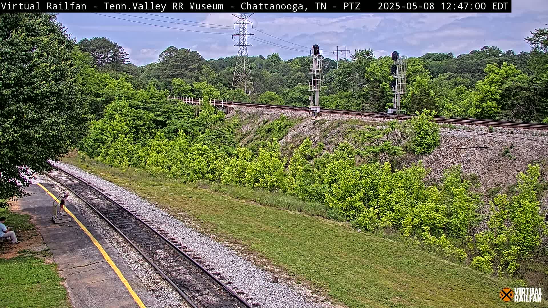 A mostly sunny view of railroad tracks running through a lush green landscape, with a person sitting on a bench near the tracks.