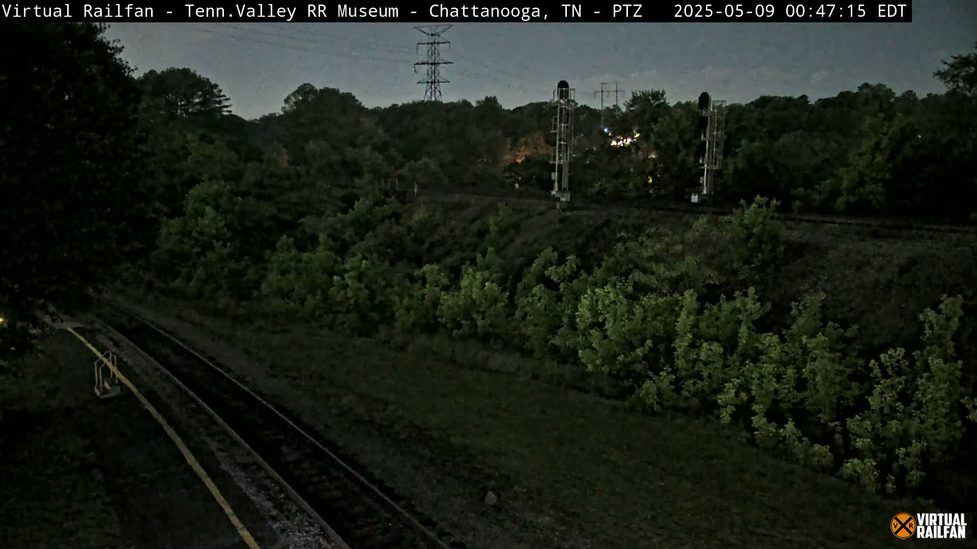 A dark, twilight view of railroad tracks running through a lush green landscape, with a tall transmission tower visible in the background.