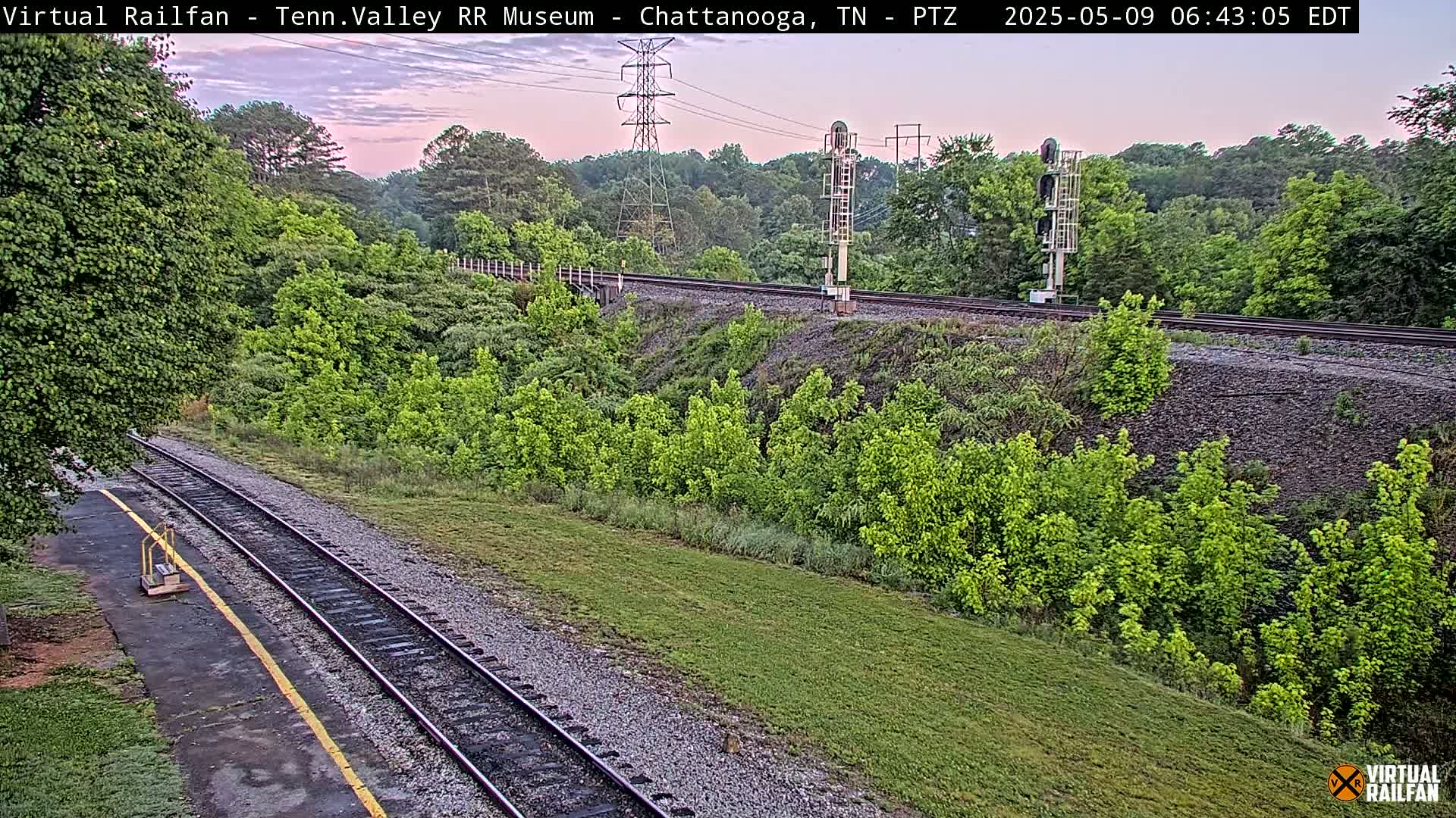 A railroad track winds through a lush green landscape under a pale pink and dusky sky.