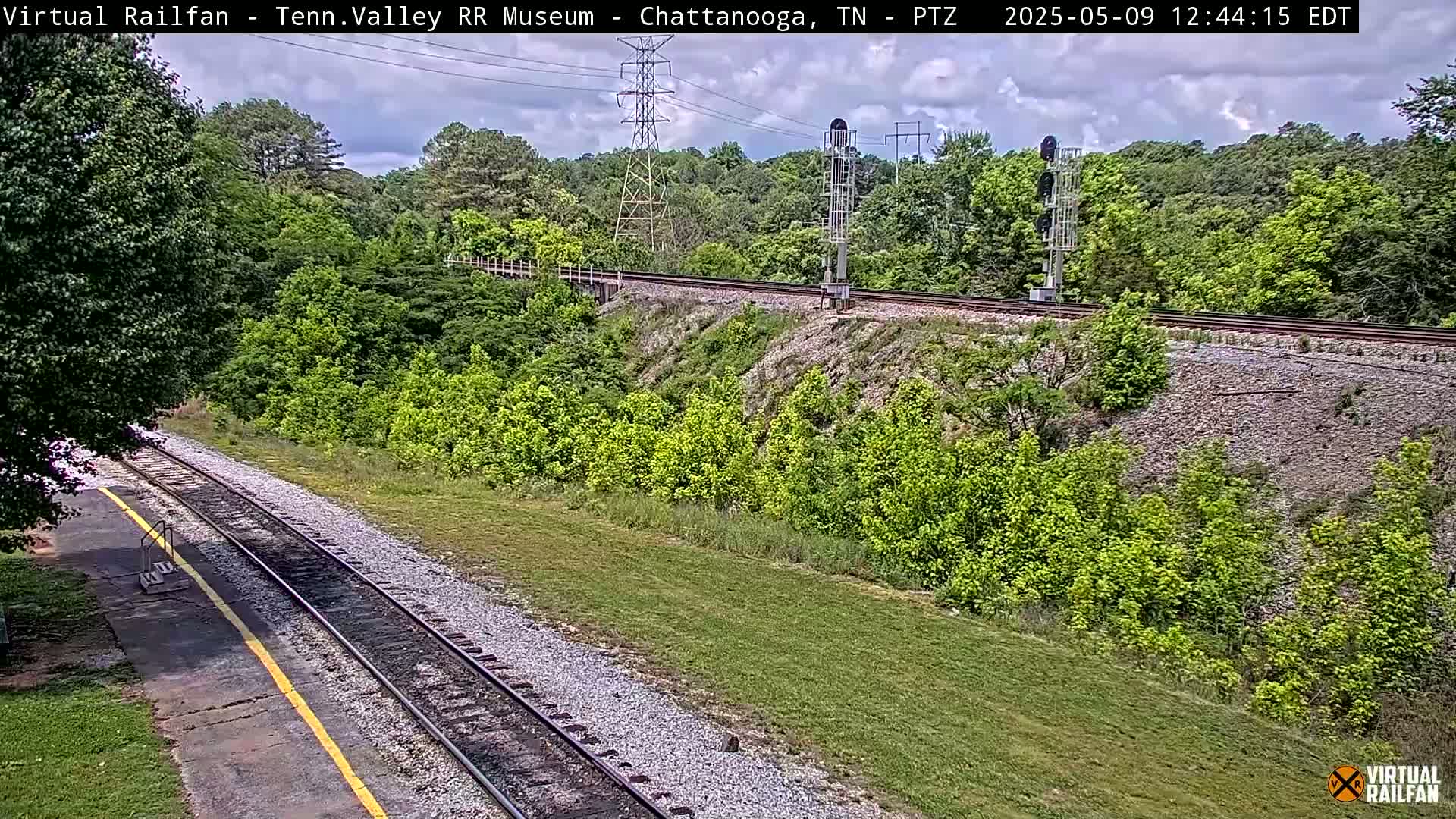 A train track curves through a lush green landscape under a partly cloudy sky.