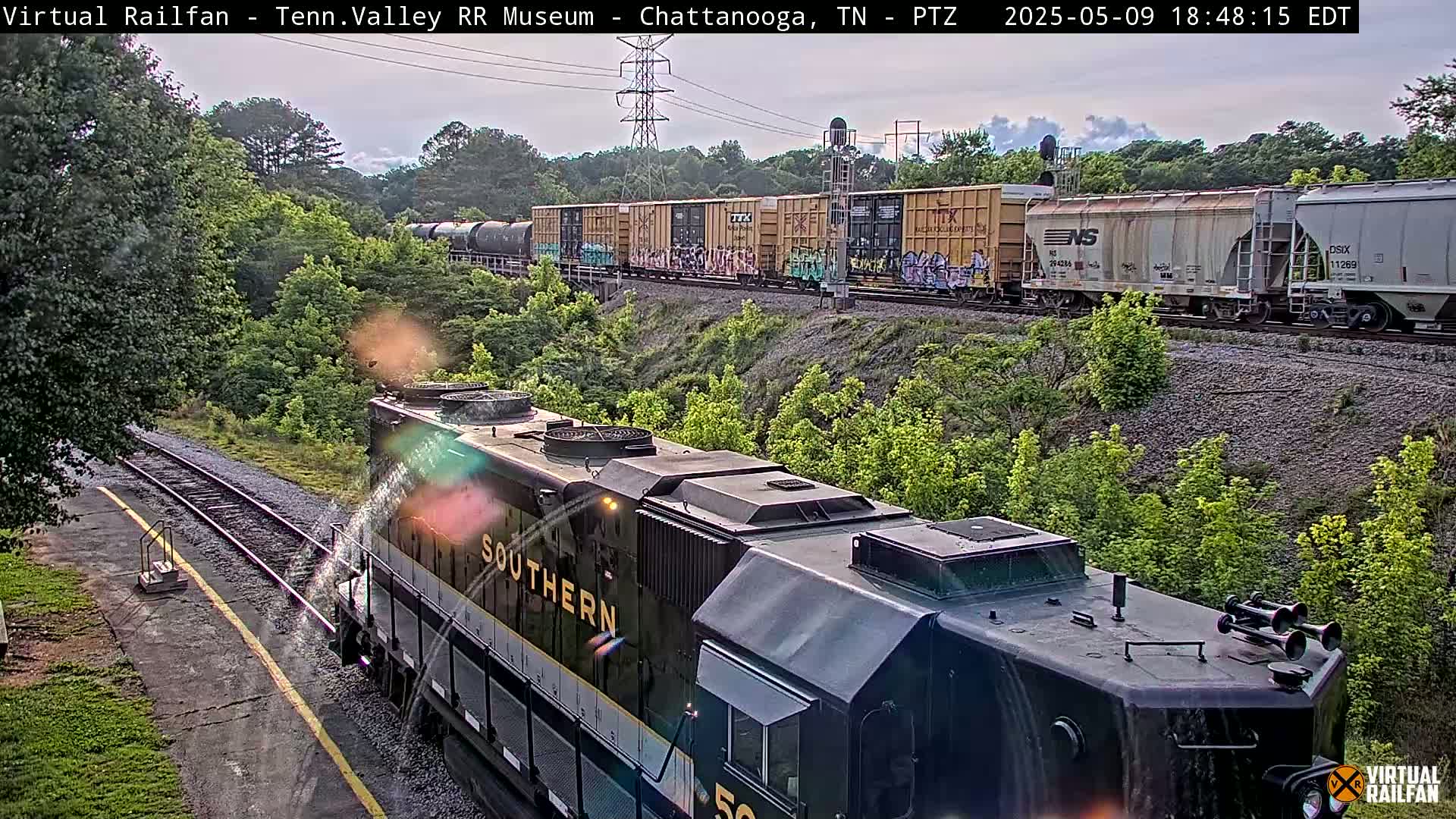 A Southern Railway locomotive approaches a railway line where a freight train, with graffiti on some of the cars, is already passing by under partly cloudy skies.