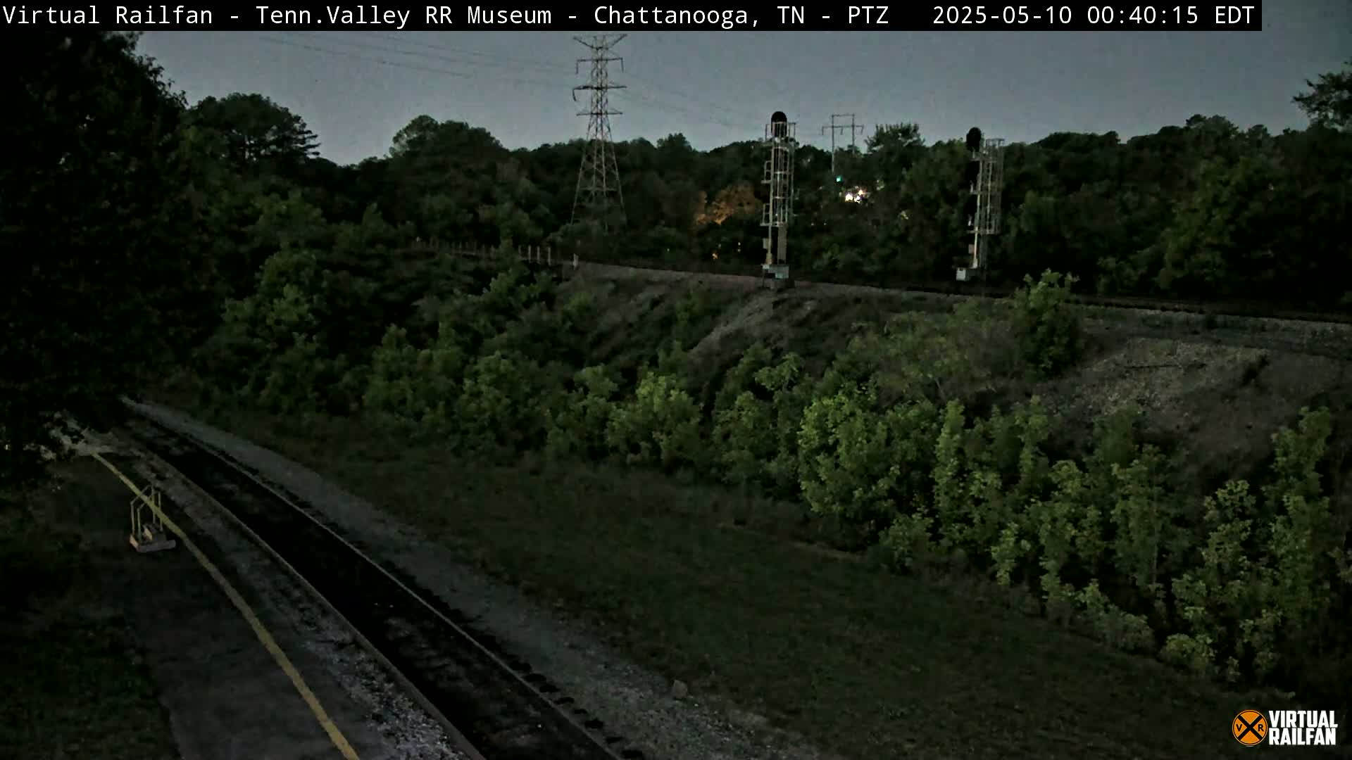 A dark, nighttime view of a railroad track running through lush green vegetation, with a tall power line and signal tower visible in the distance under a dusky sky.