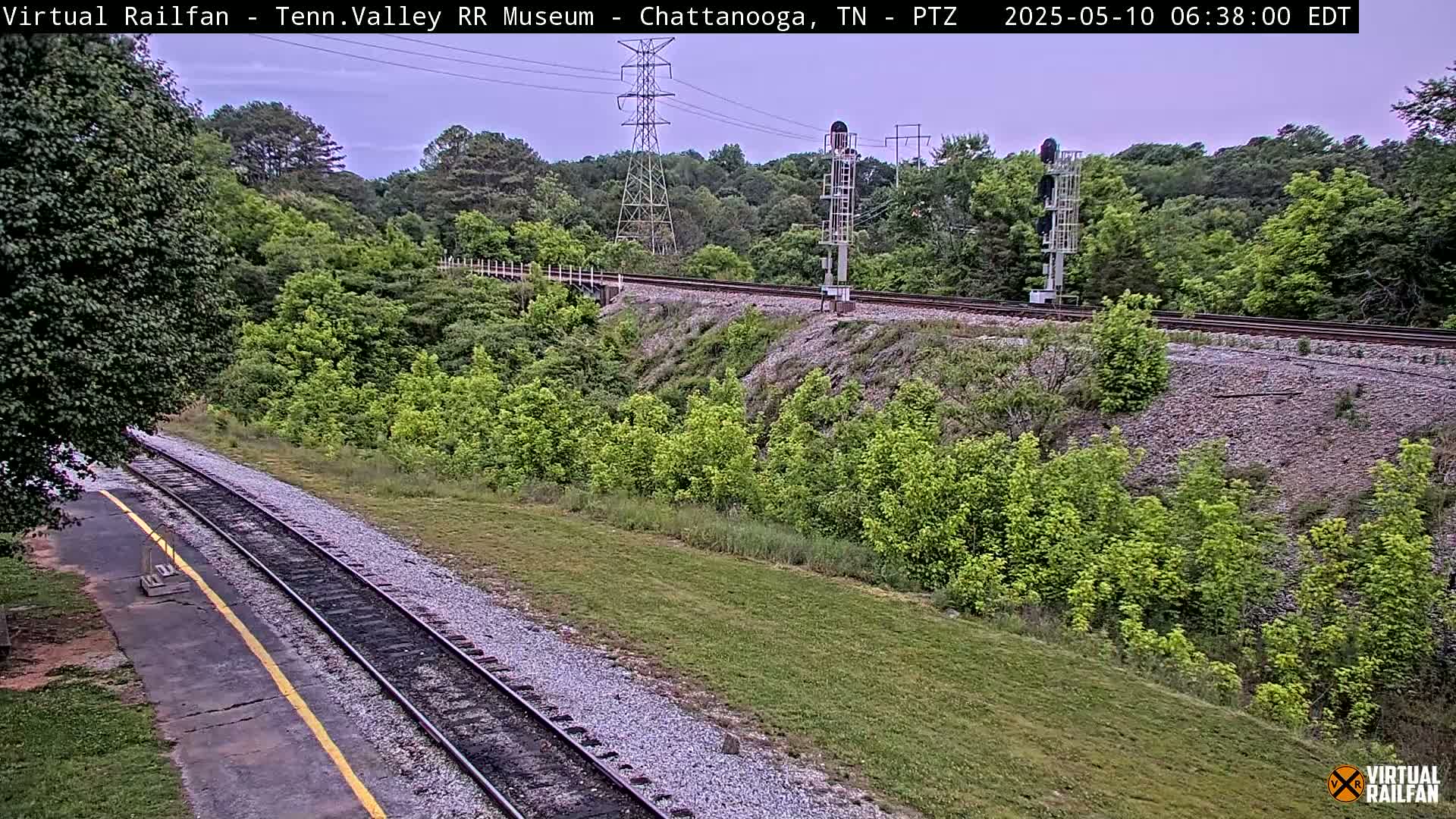 A high-angle, long shot of a railway track winding through a lush green landscape under a partly cloudy sky.