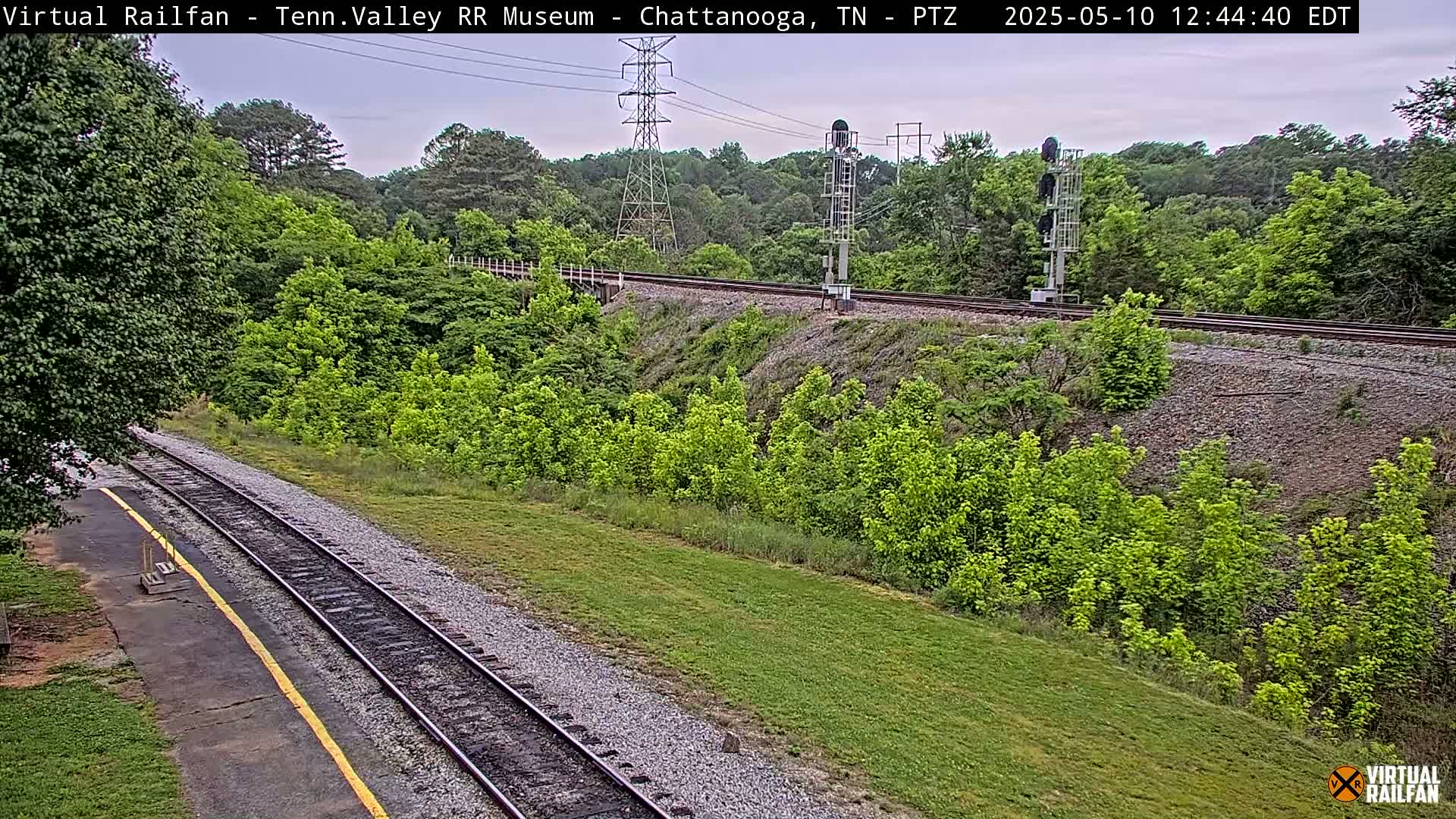 A mostly sunny view of railroad tracks cutting through a lush green landscape, with power lines and a signal tower visible in the background.