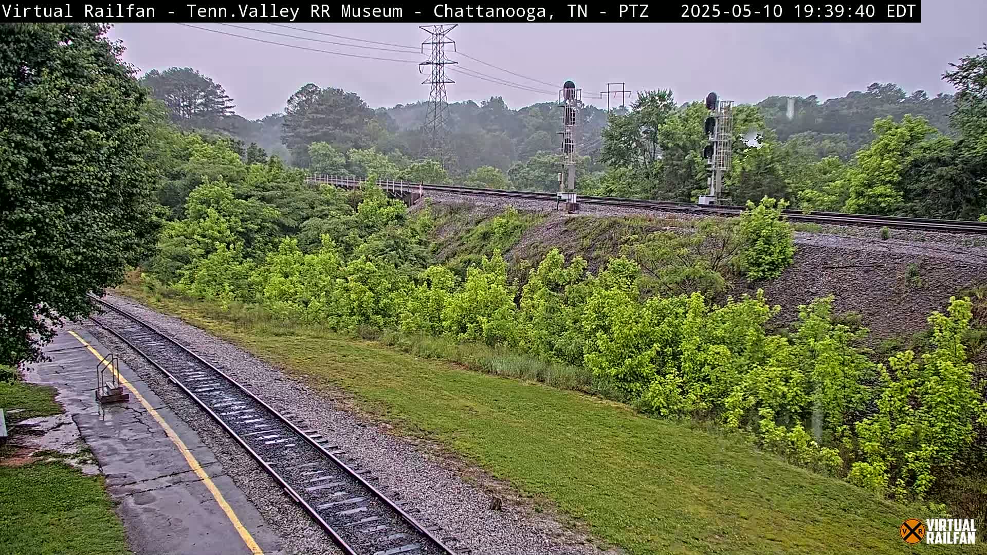 A rain-slicked railroad track winds through a lush green landscape under an overcast sky.