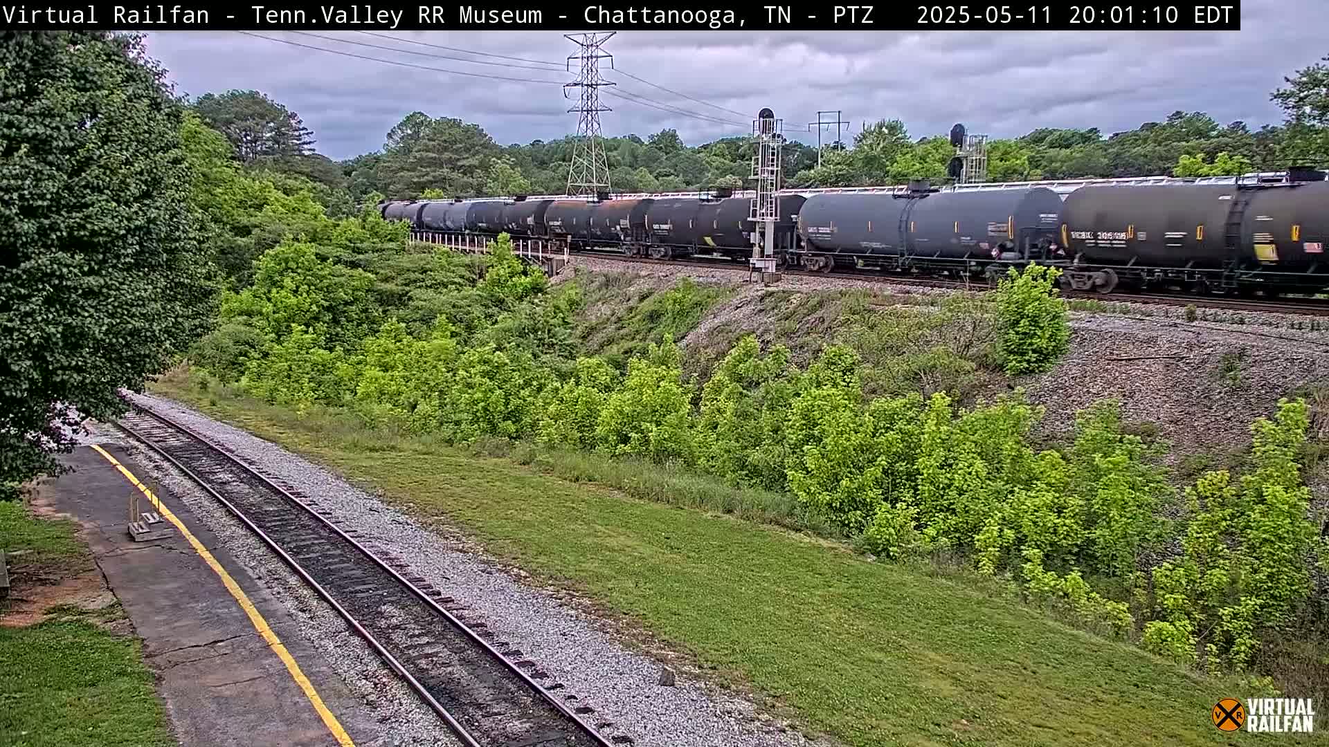 A long freight train of dark tanker cars passes by a grassy area with trees under a partly cloudy sky.