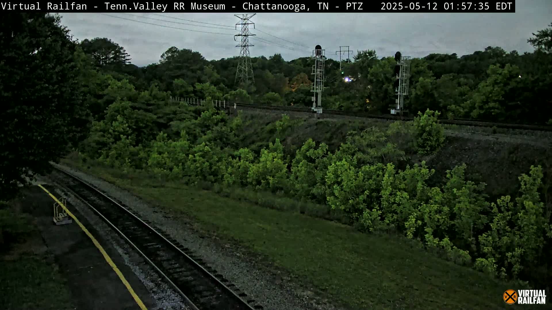 A high-angle, long shot shows a railroad track winding through lush green vegetation under a gray, overcast sky.