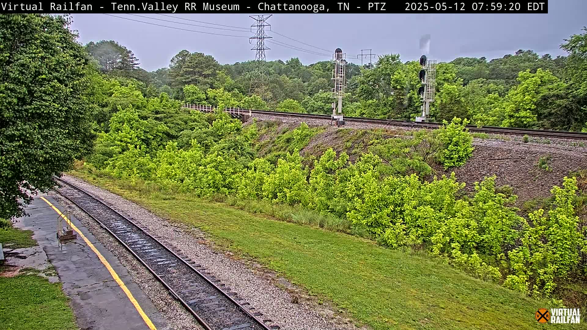 A railroad track winds through a lush green landscape under an overcast sky, passing by a signal tower and a powerline.