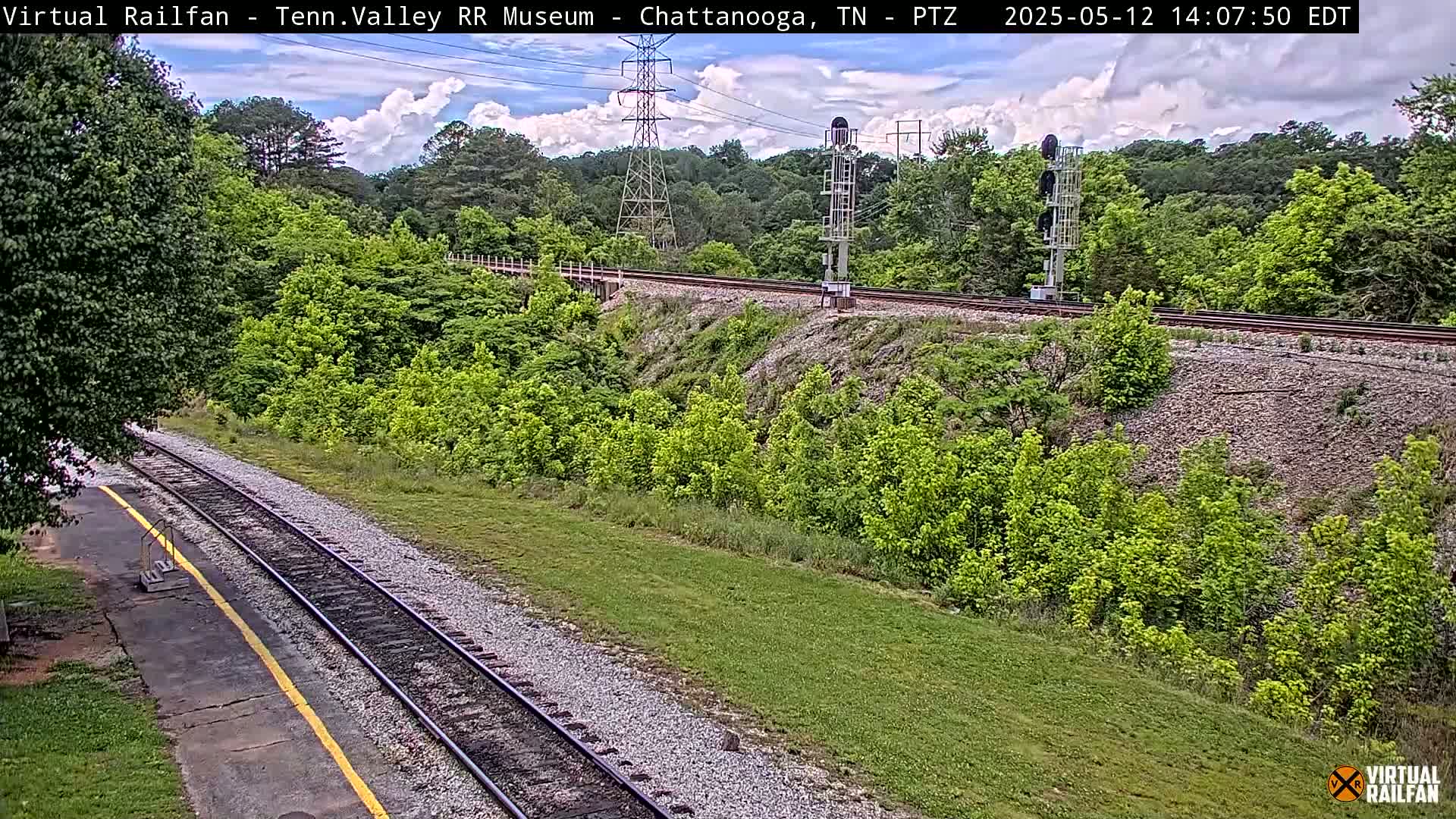 A partly cloudy day shows a railroad track winding through a lush green landscape, with a signal tower visible in the middle ground.