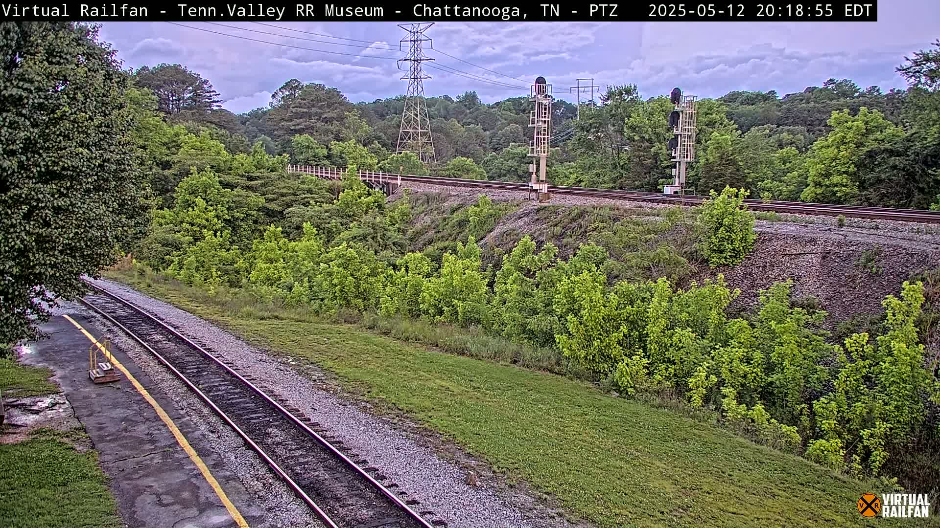 A railroad track cuts through a lush green landscape under a partly cloudy sky.