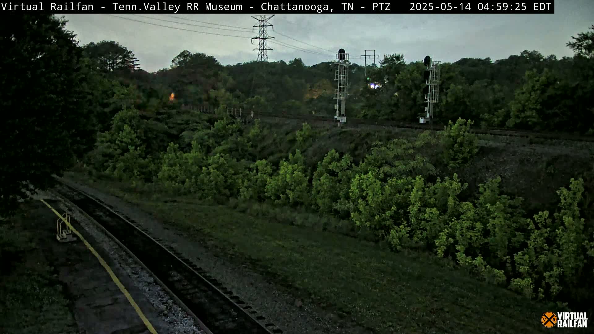 A high-angle, long shot shows a railway track winding through a lush green landscape under an overcast sky.