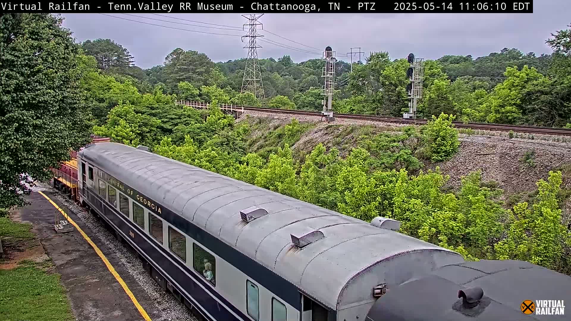 A long, silver passenger train car, labeled "Central of Georgia," is parked on a railway track next to lush green foliage under an overcast sky.