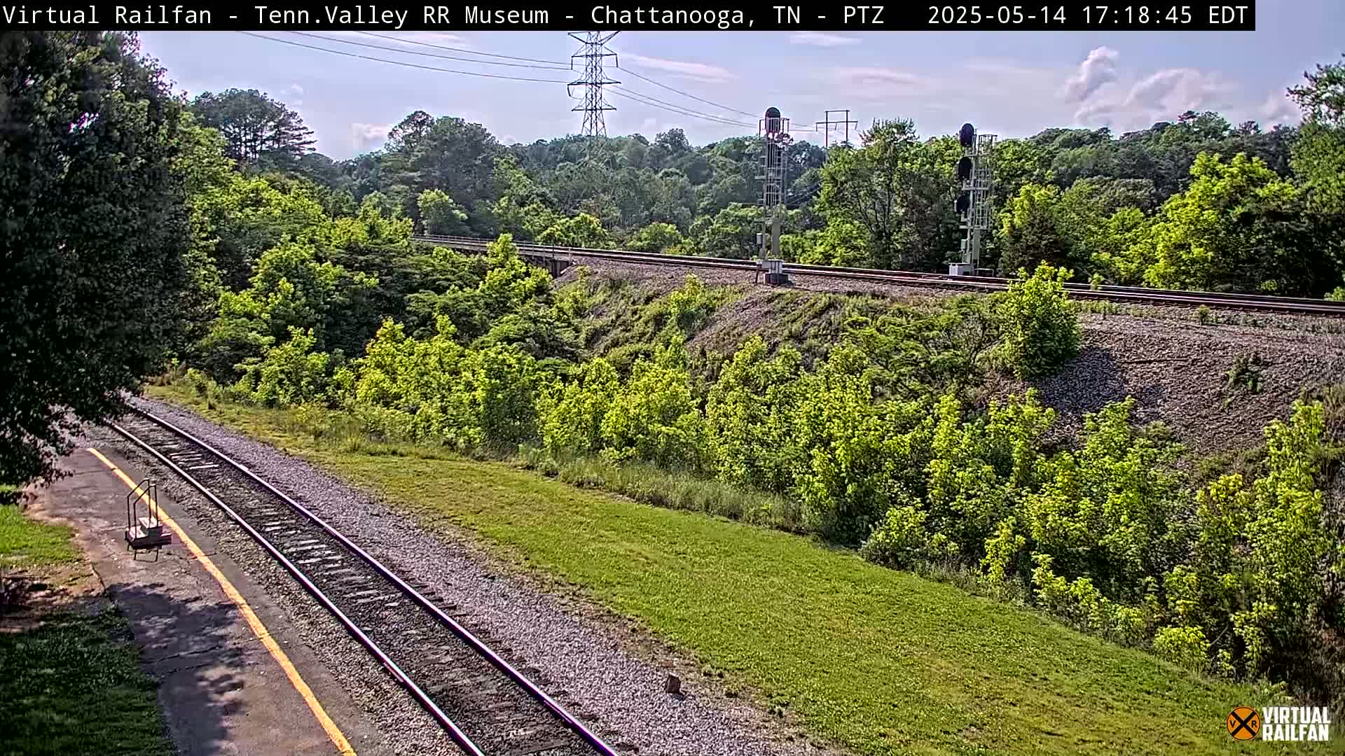 A partly sunny view of a train track winding through a lush green landscape, with a signal tower visible in the distance.