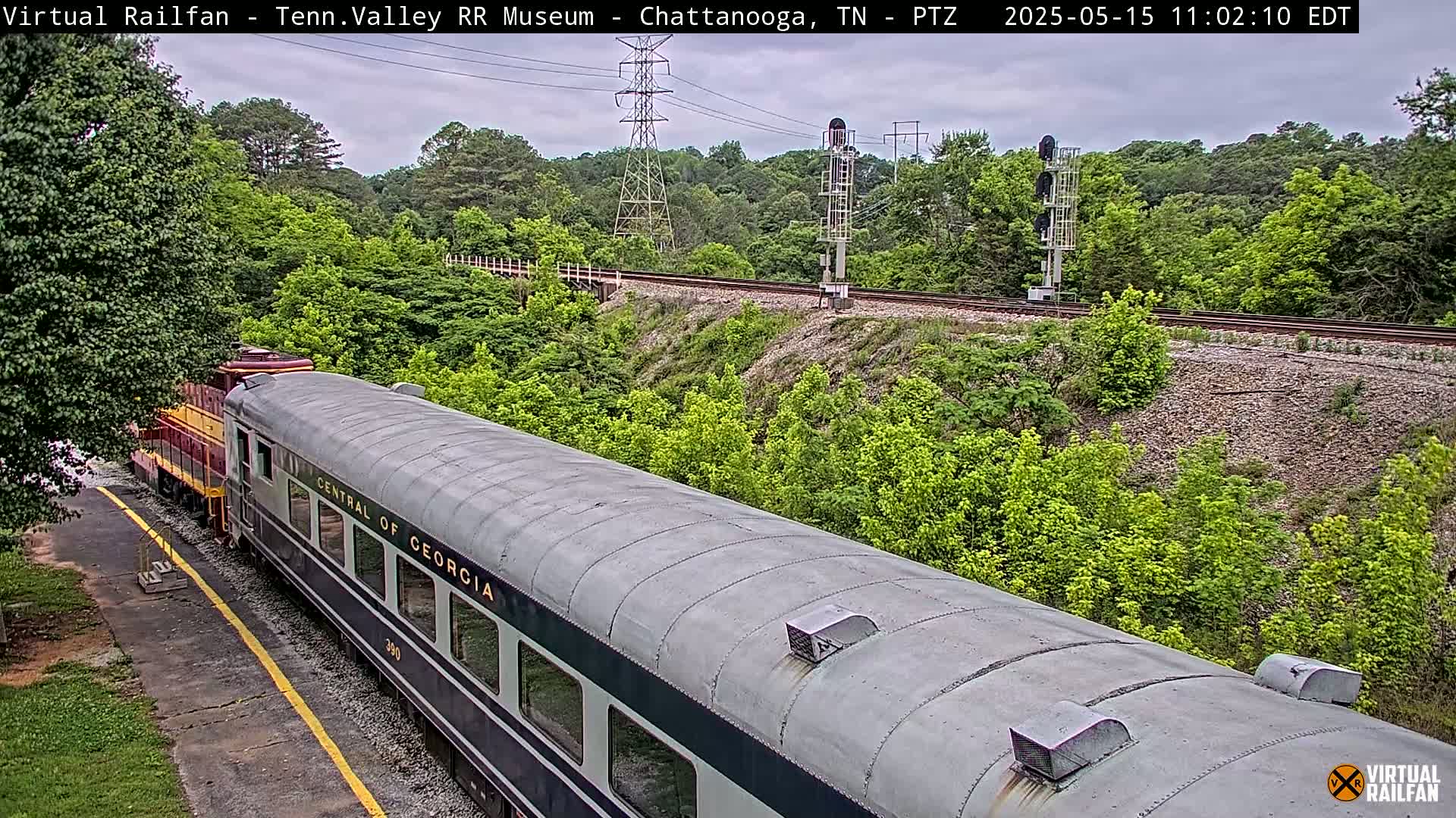 A passenger train car, labeled "Central of Georgia," is parked next to a railway track that runs through a lush green landscape under a cloudy sky.