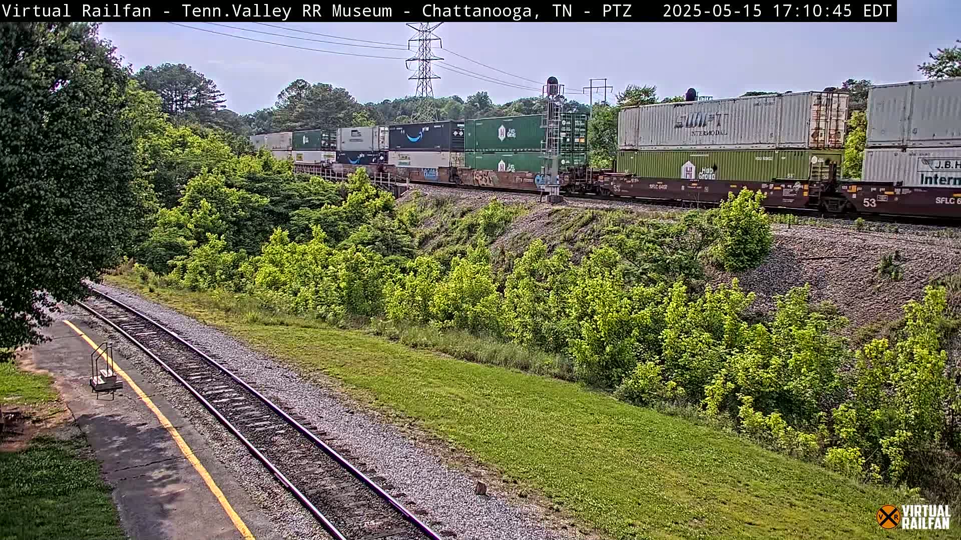 A freight train carrying multiple shipping containers passes by a grassy area next to train tracks under a partly cloudy sky.