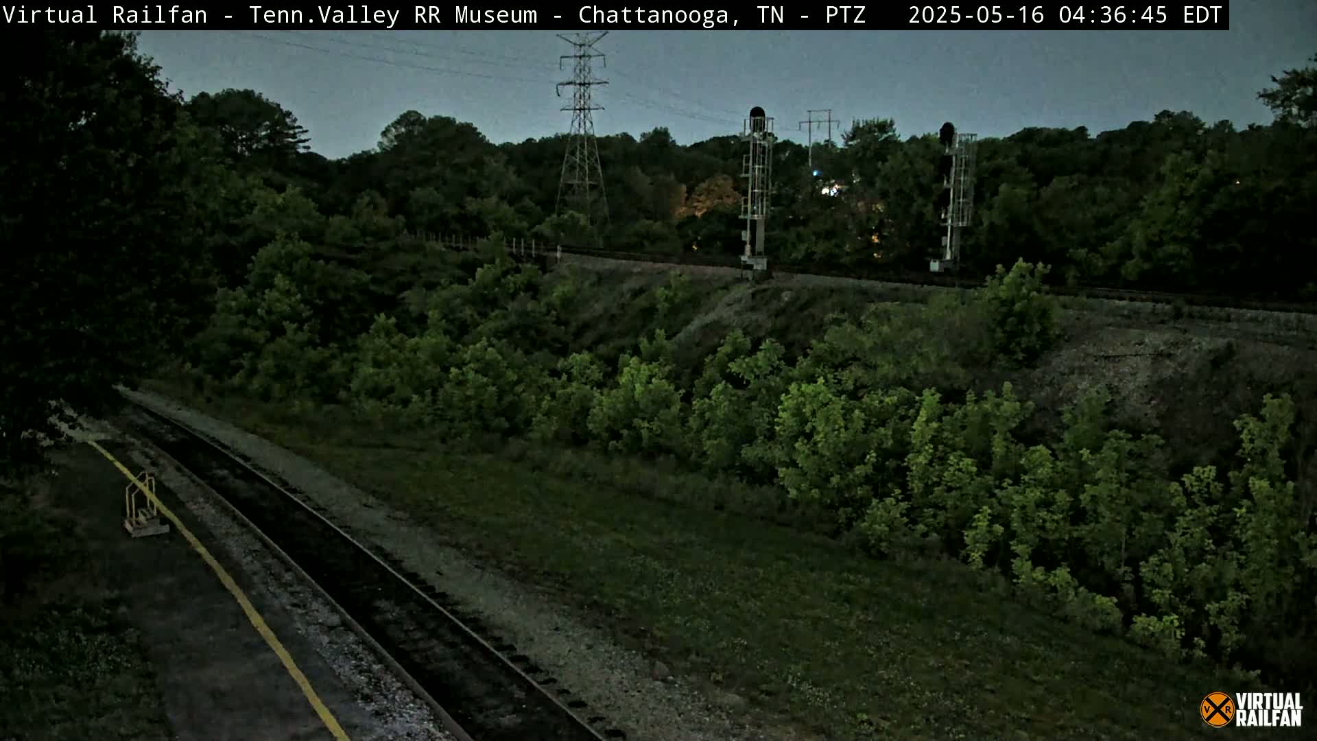 A high-angle, twilight view shows a train track winding through a lush, green landscape, with tall power lines and signal equipment visible in the background.