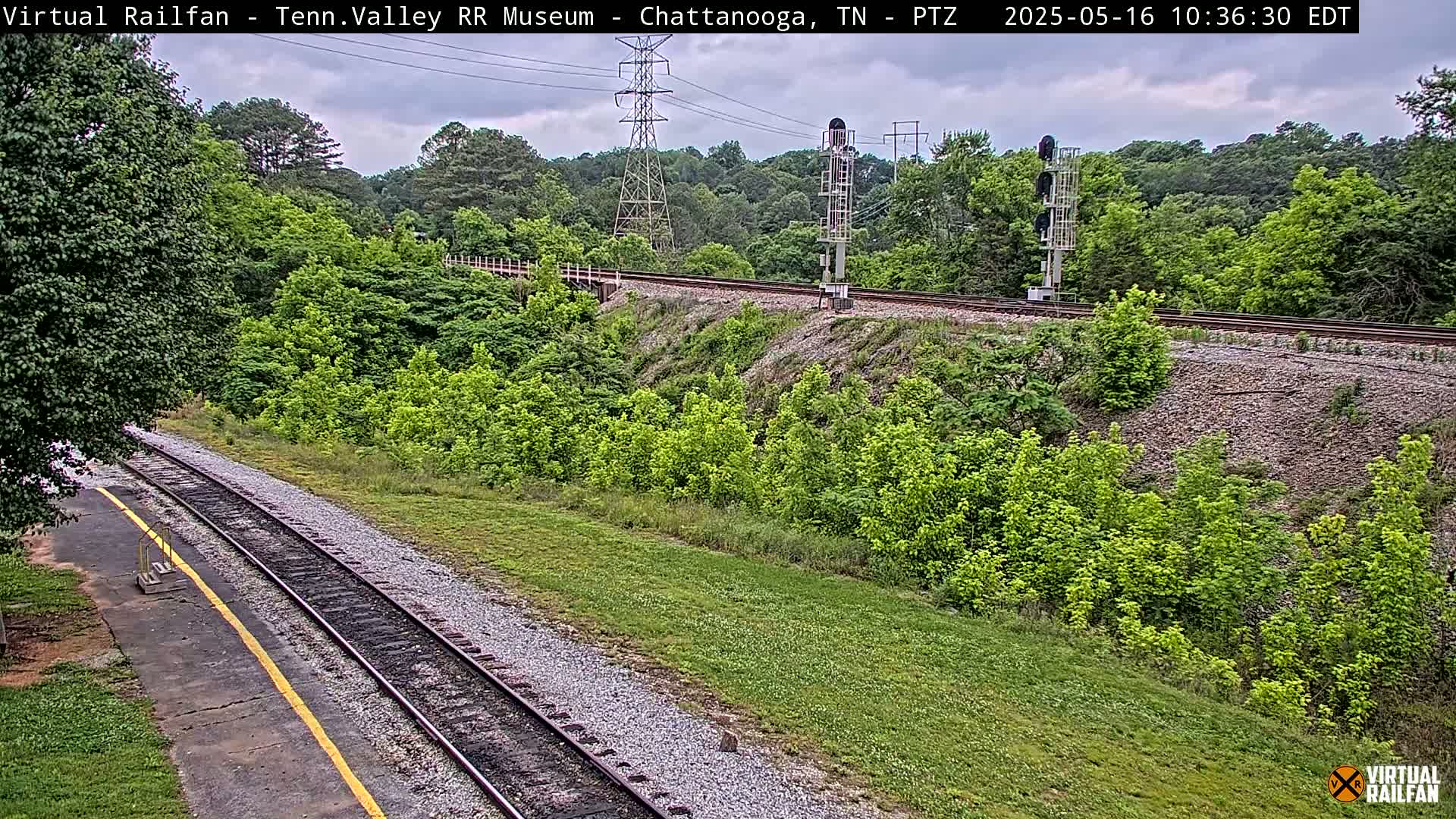 A railroad track runs through a grassy area alongside a lush green hillside under a partly cloudy sky.