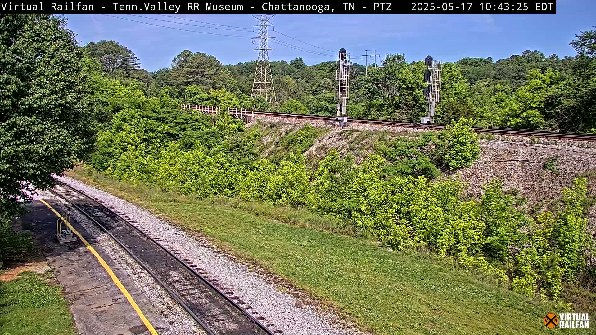 A sunny day shows a view of railroad tracks that wind through a lush green landscape, alongside a tall electrical tower and signal lights.