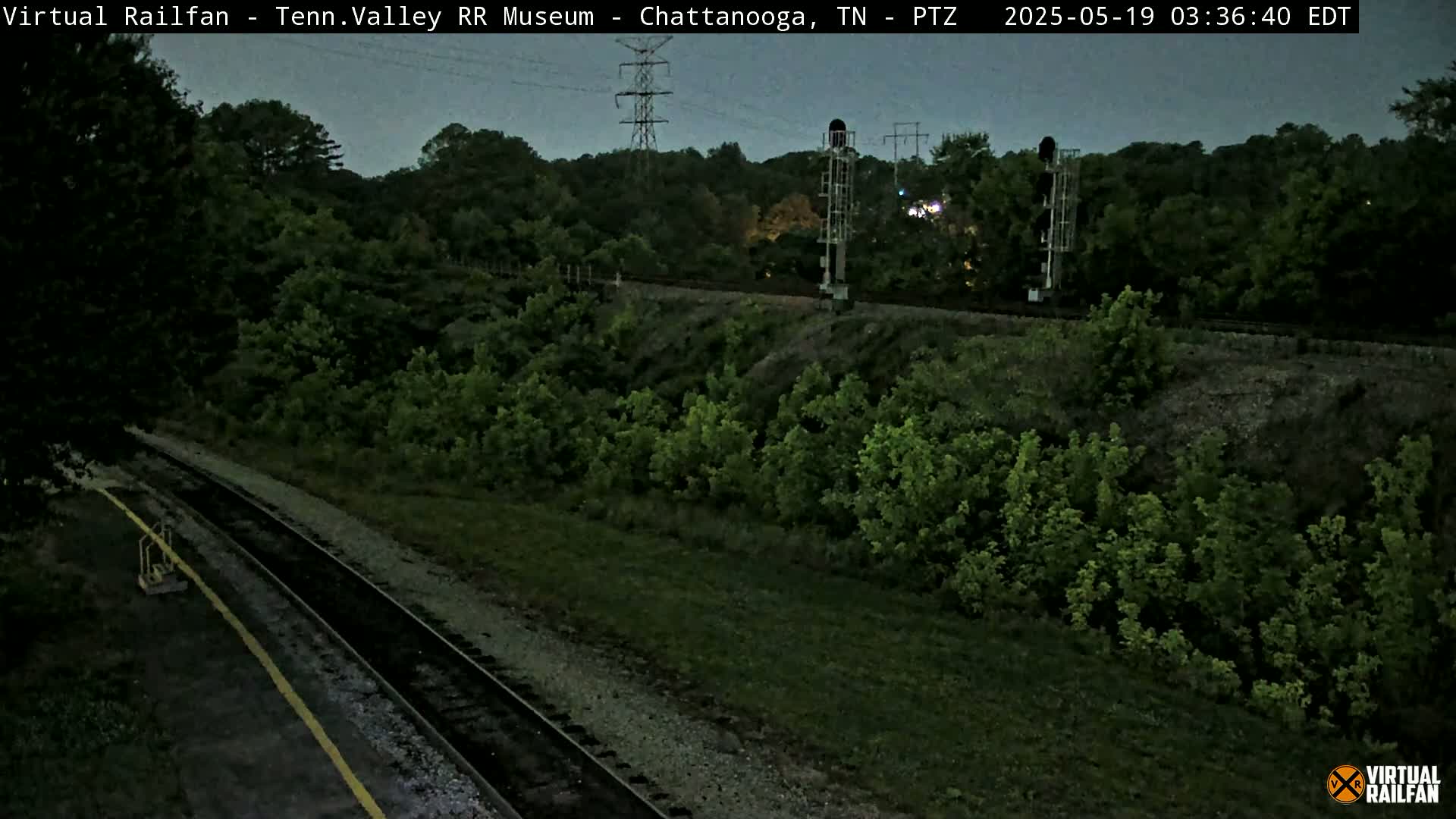 A dark-green, lush landscape surrounds a set of train tracks at twilight, with power lines visible in the background.