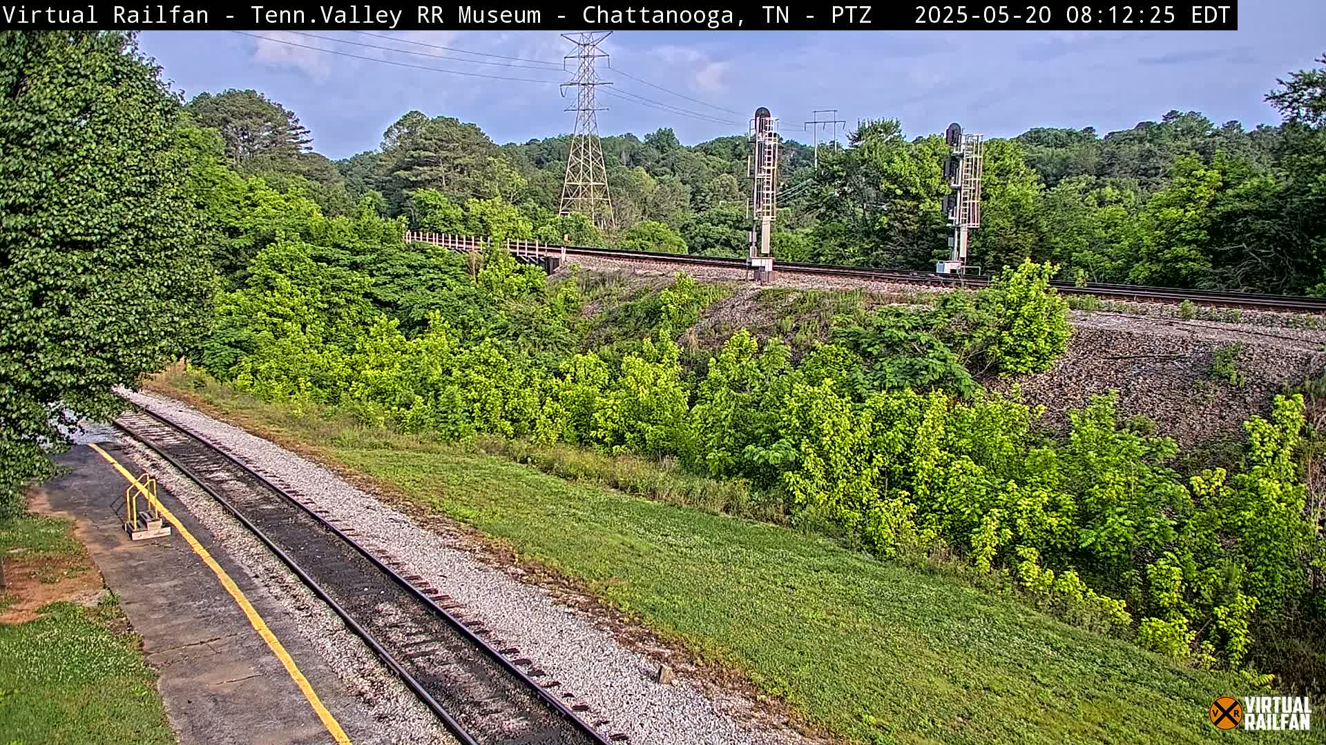 A partly sunny view of railroad tracks winding through a lush green landscape, with a tall transmission tower in the background.