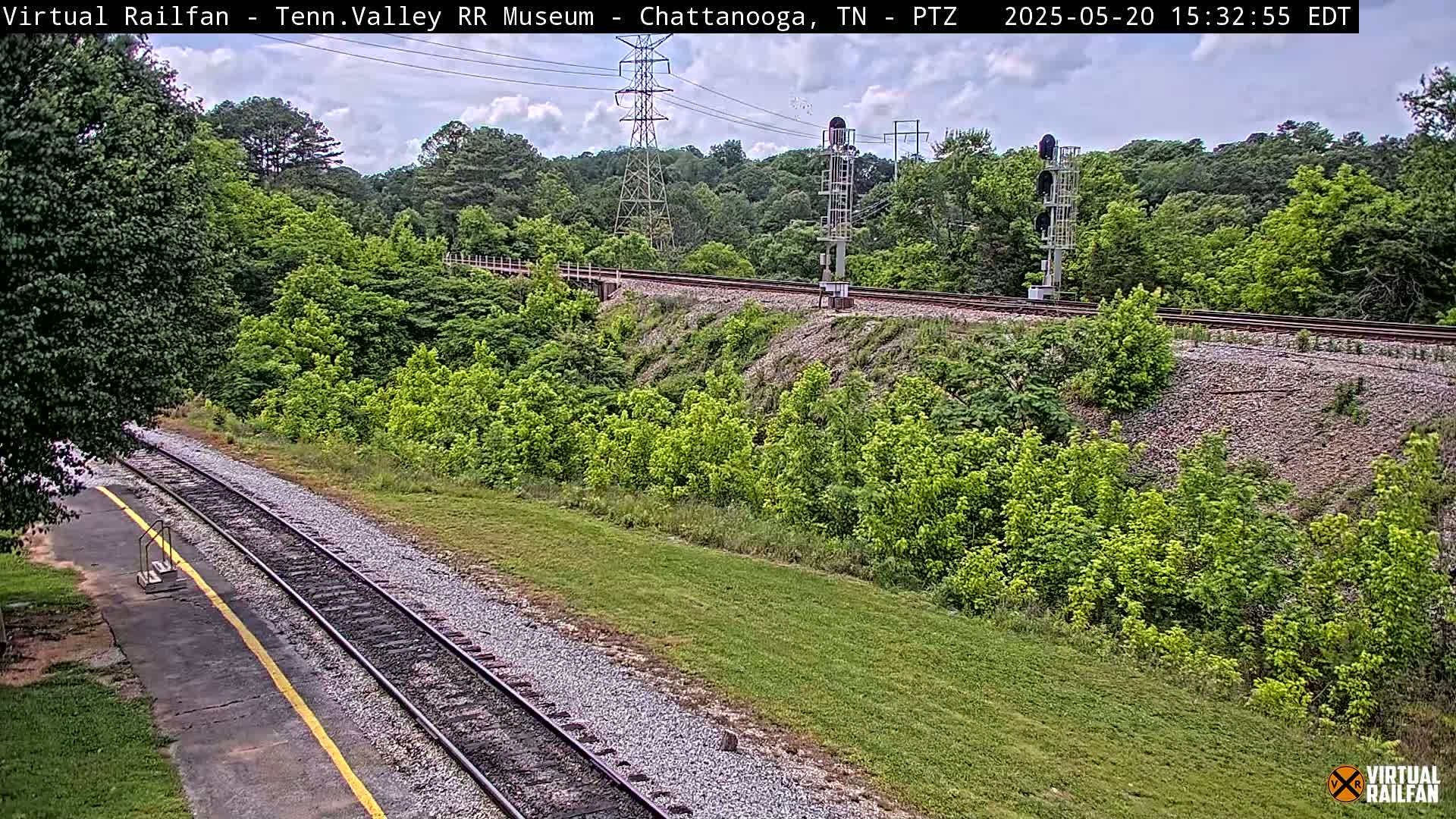 A partly cloudy day shows a view of railroad tracks winding through a lush green landscape, with a small platform visible in the foreground.
