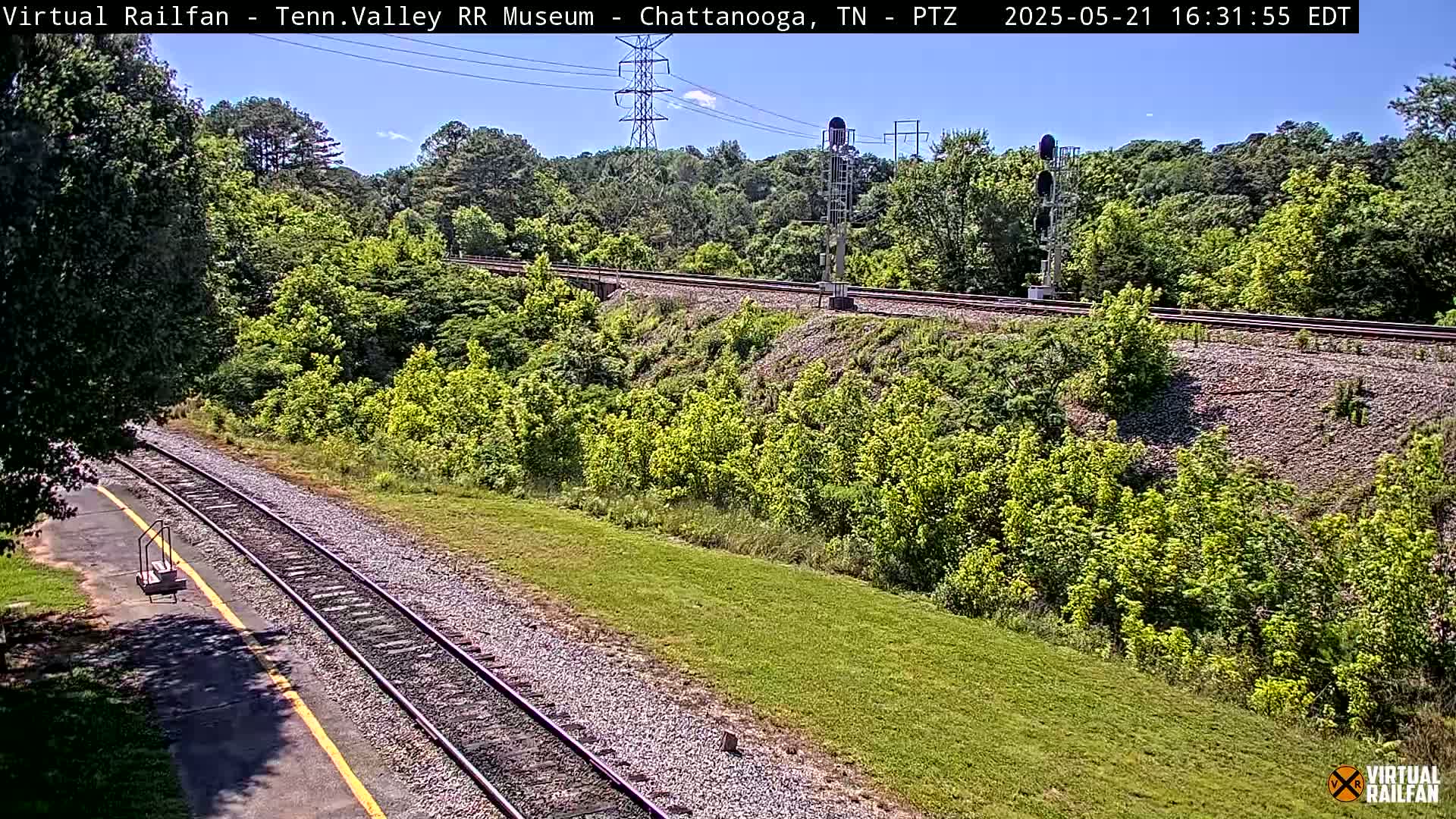 A sunny day reveals a railway track winding through a lush green landscape, bordered by trees and a grassy embankment.