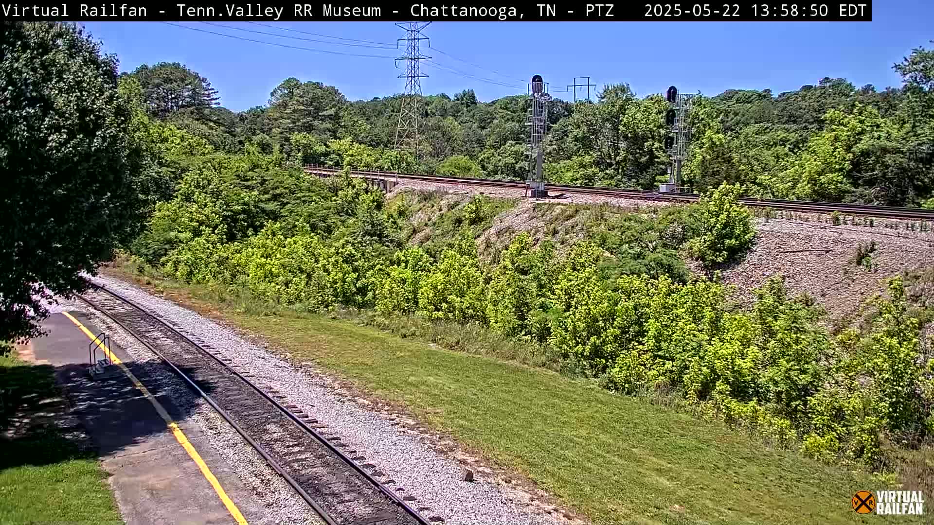 A sunny view of a railway track winding through a green landscape, with trees and shrubs lining the sides and a tall structure visible in the distance.