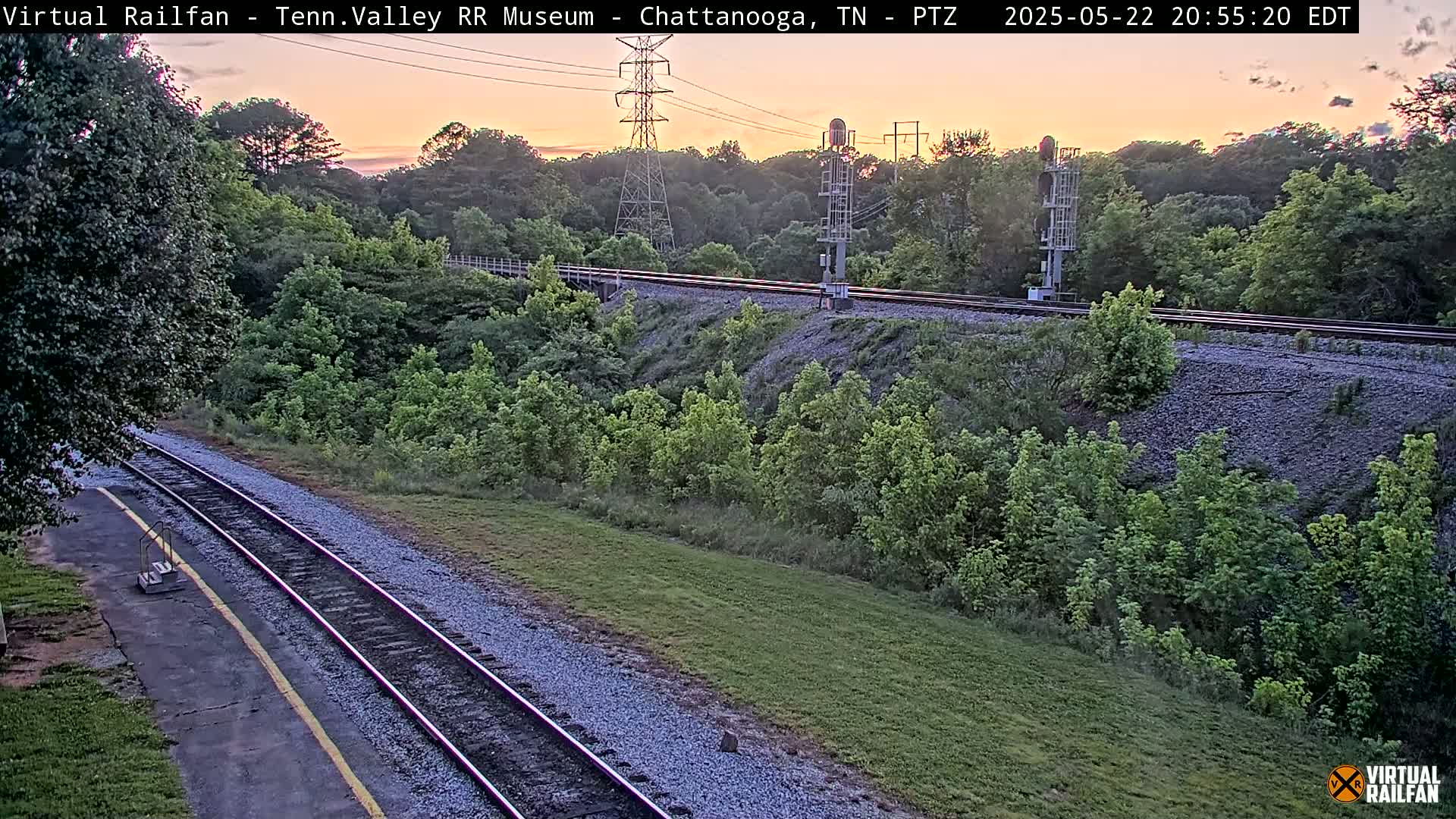 A sunset view of a railway track winding through a grassy area with lush green vegetation and a tall transmission tower in the background.