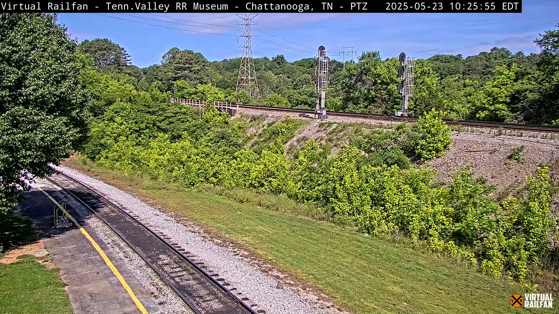 A sunny day overlooking a railway track that runs through a lush green landscape, with trees and shrubs lining both sides of the tracks.
