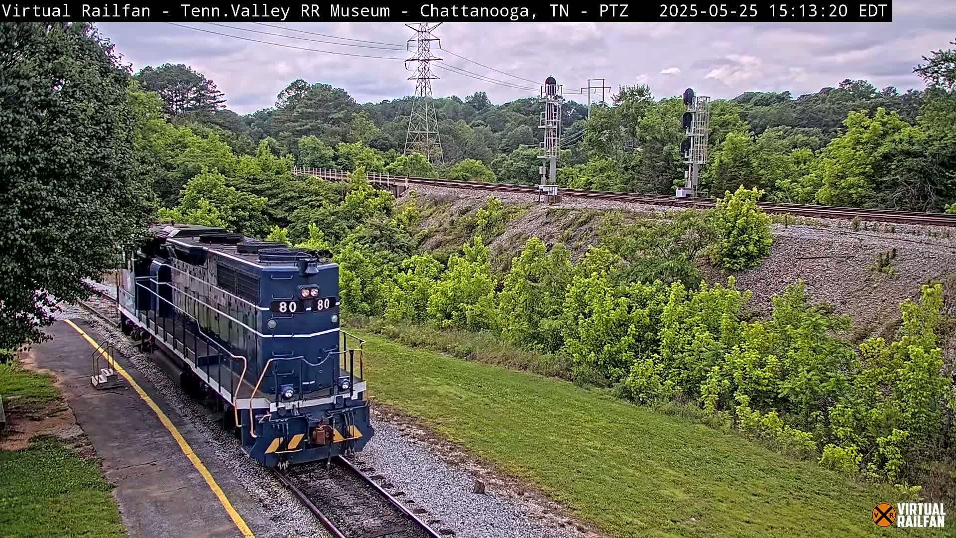 A blue diesel locomotive is traveling along a railway track, surrounded by lush green vegetation under a partly cloudy sky.