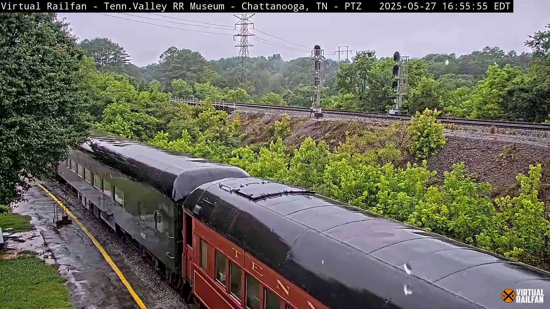 A long, dark-colored passenger train is shown moving along railroad tracks next to lush green vegetation on a rainy day.