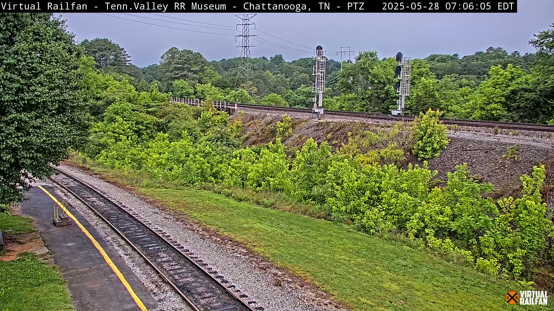 A mostly clear day shows a view of train tracks that wind through a lush green landscape with a small section of paved area and a tall powerline.